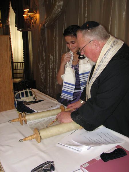 A person in a tallit and kippah assisting a person near a Torah scroll.