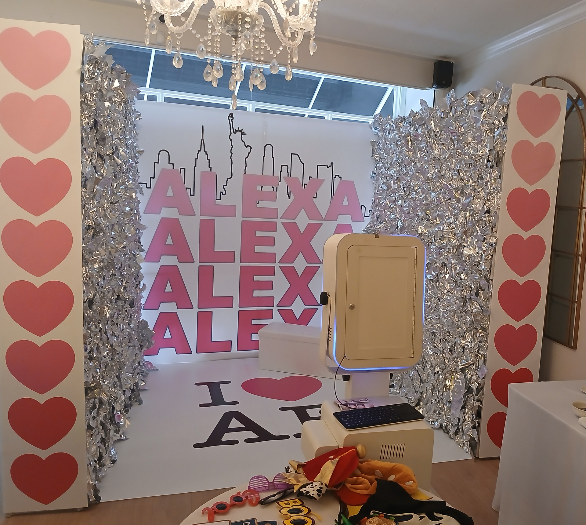 Photo booth setup with silver fringe backdrop, pink heart decorations, and