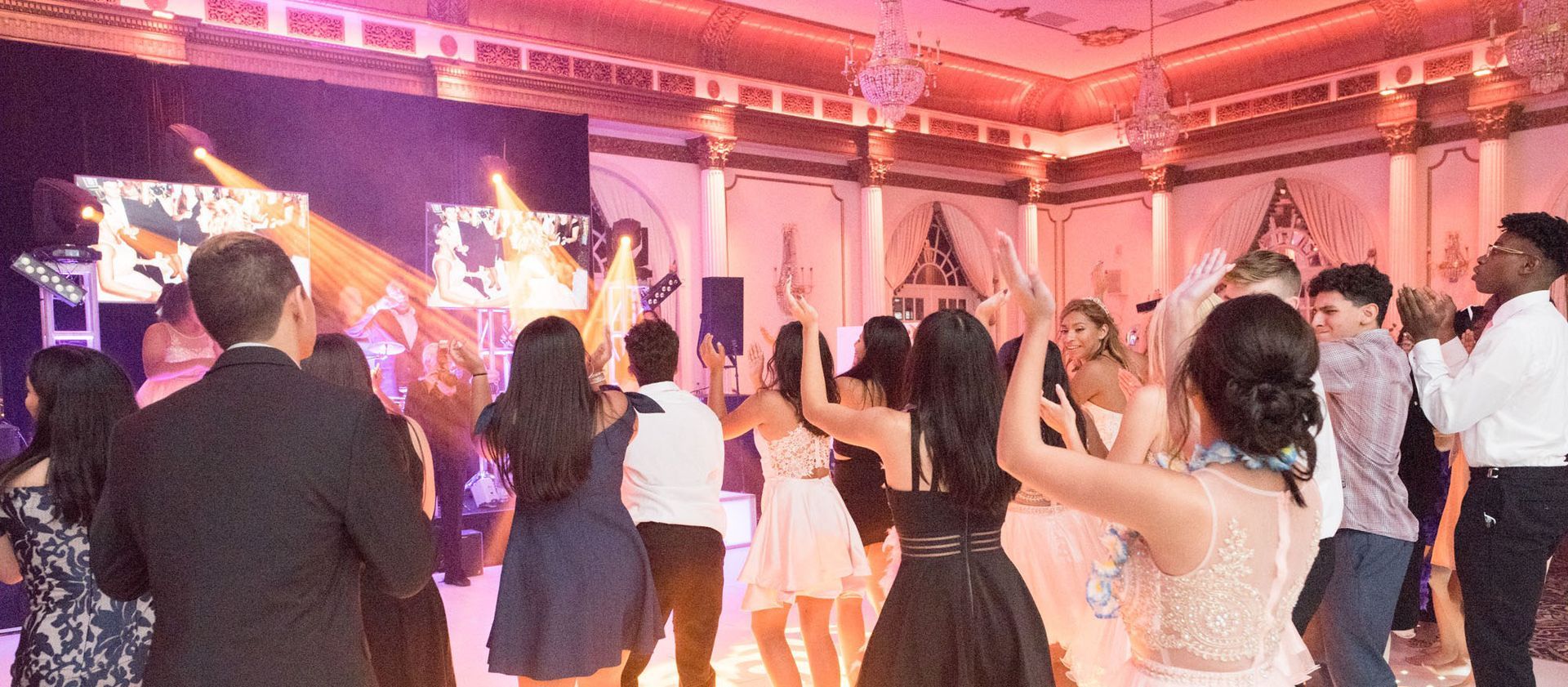 People dancing in a brightly lit ballroom. A DJ booth is visible.