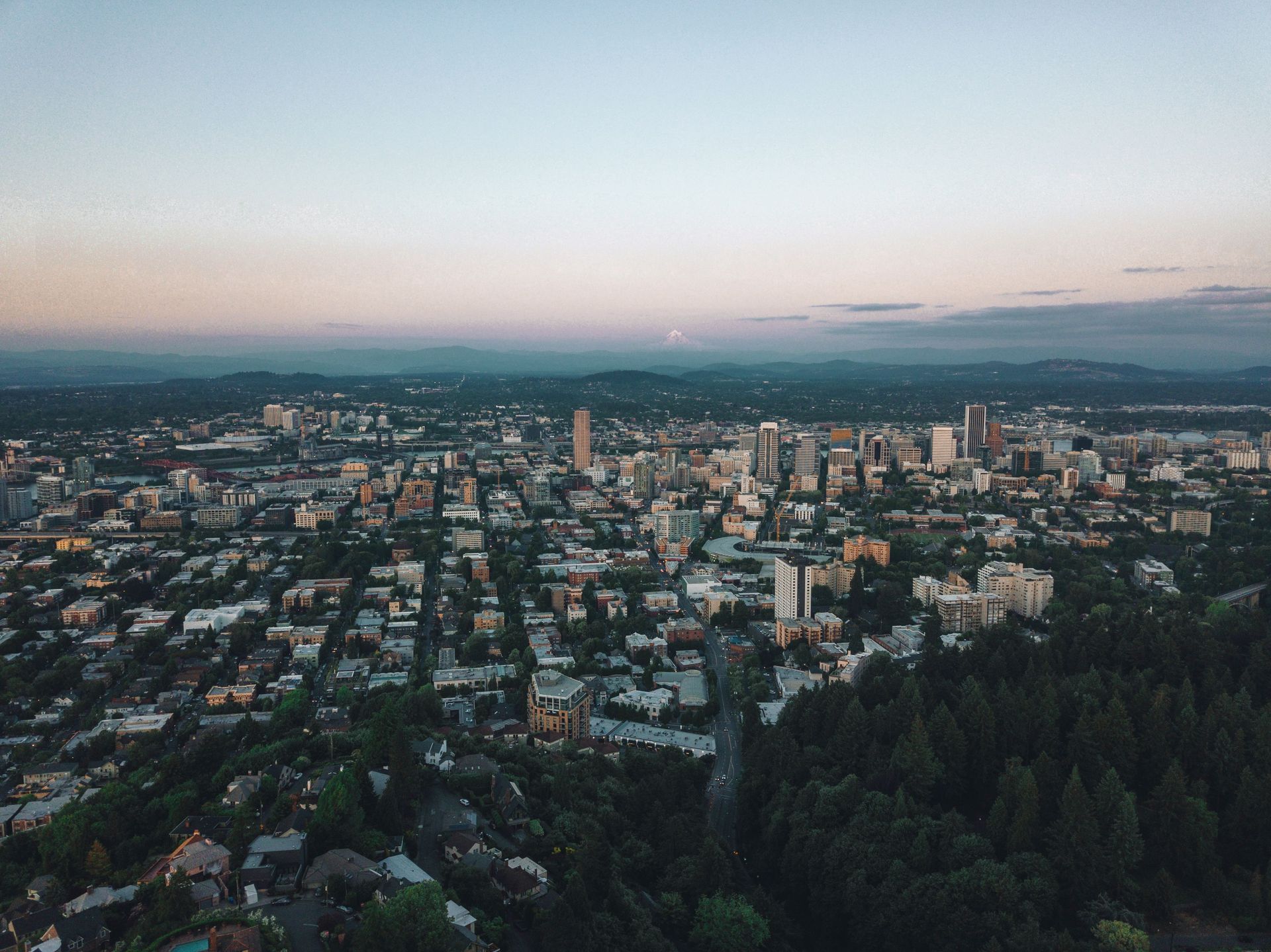 Aerial view of a city at dusk with buildings, streets, and a dark forested area in the foreground