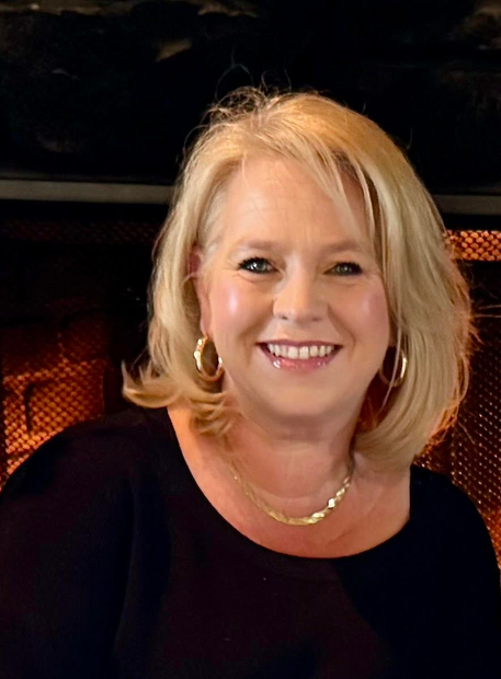 about us Smiling person with blonde hair wearing a black top and gold necklace, posed in front of a warm fireplace.