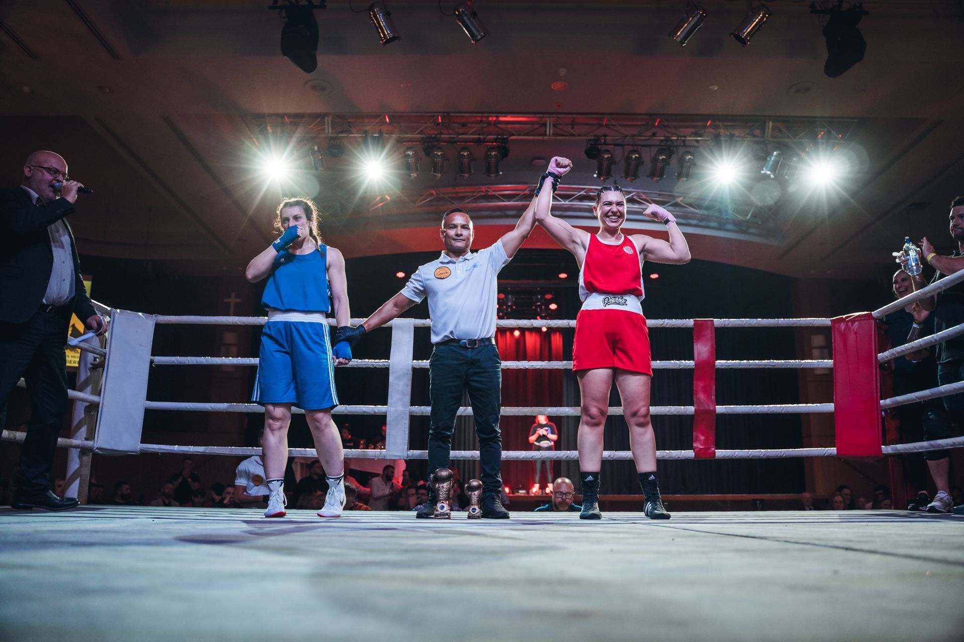 Boxer in red, arms raised in victory, referee raising her arm. Other boxer in blue looks on. Boxing ring.