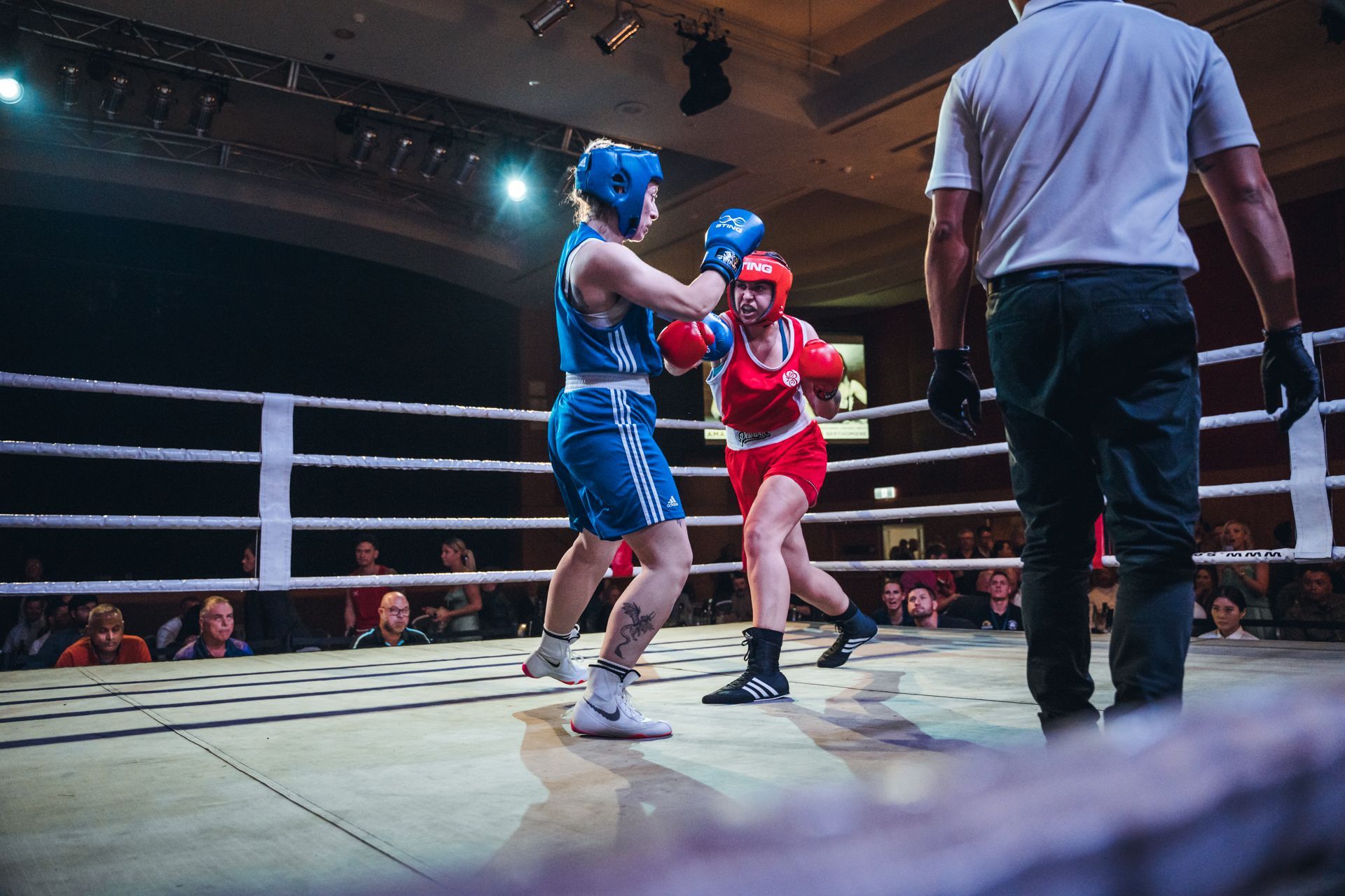 Two female boxers in a ring; one in blue, one in red, with a referee watching, surrounded by spectators.