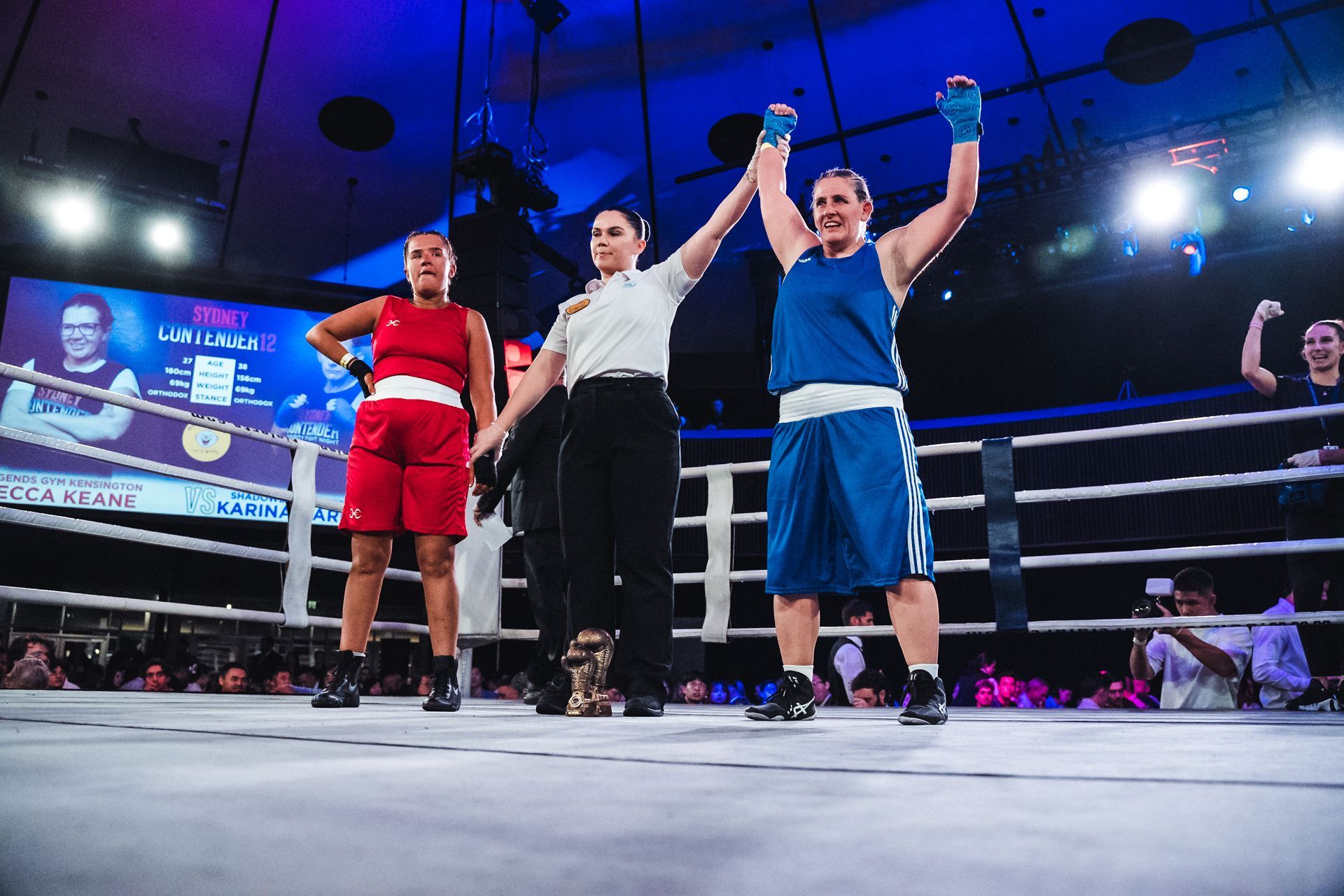 Boxer in blue, arms raised in victory, referee in center, opponent in red, boxing ring setting.