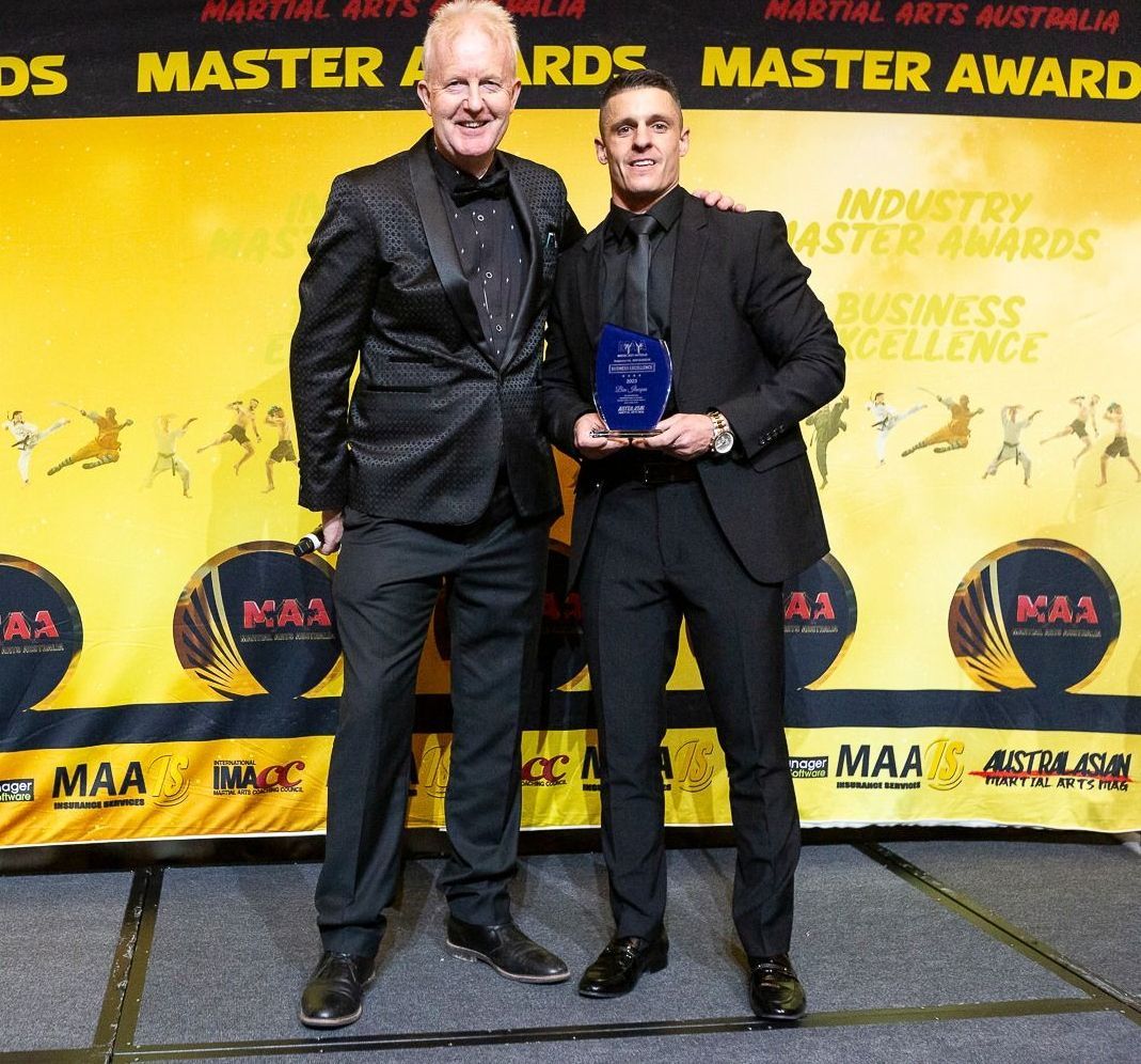 Two men in suits at an awards event; one holds a trophy. Yellow background with event logos and text.