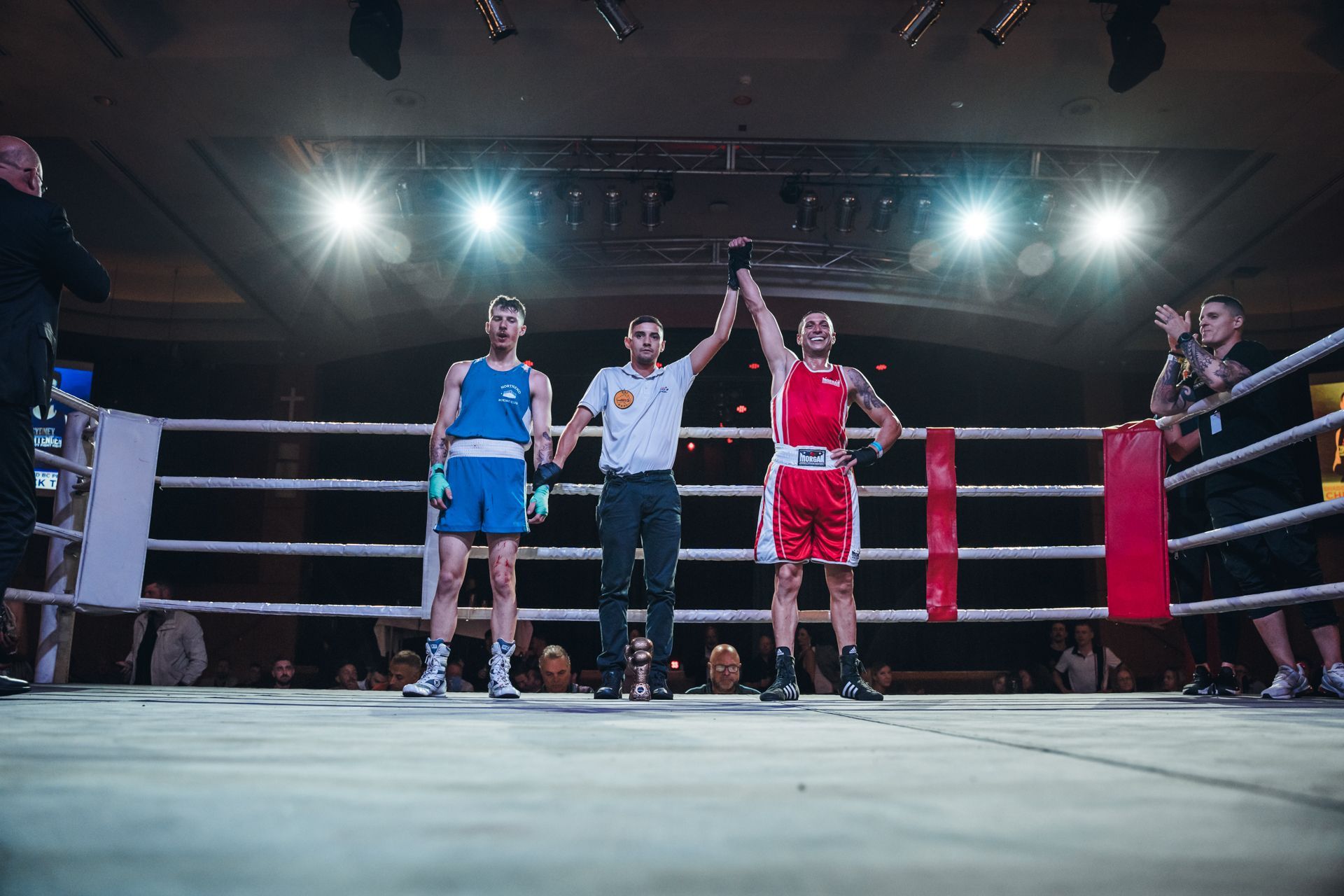 Boxer in red wins match, referee raises his arm in a boxing ring, blue-clad boxer stands nearby.