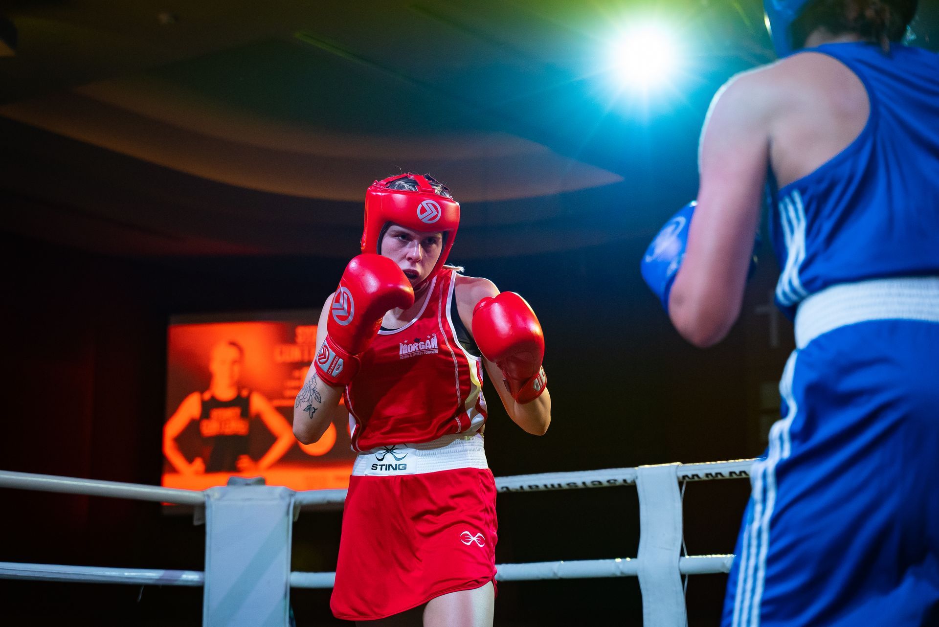 Boxer in red gear faces opponent in blue, inside a boxing ring. Bright lights illuminate the match.