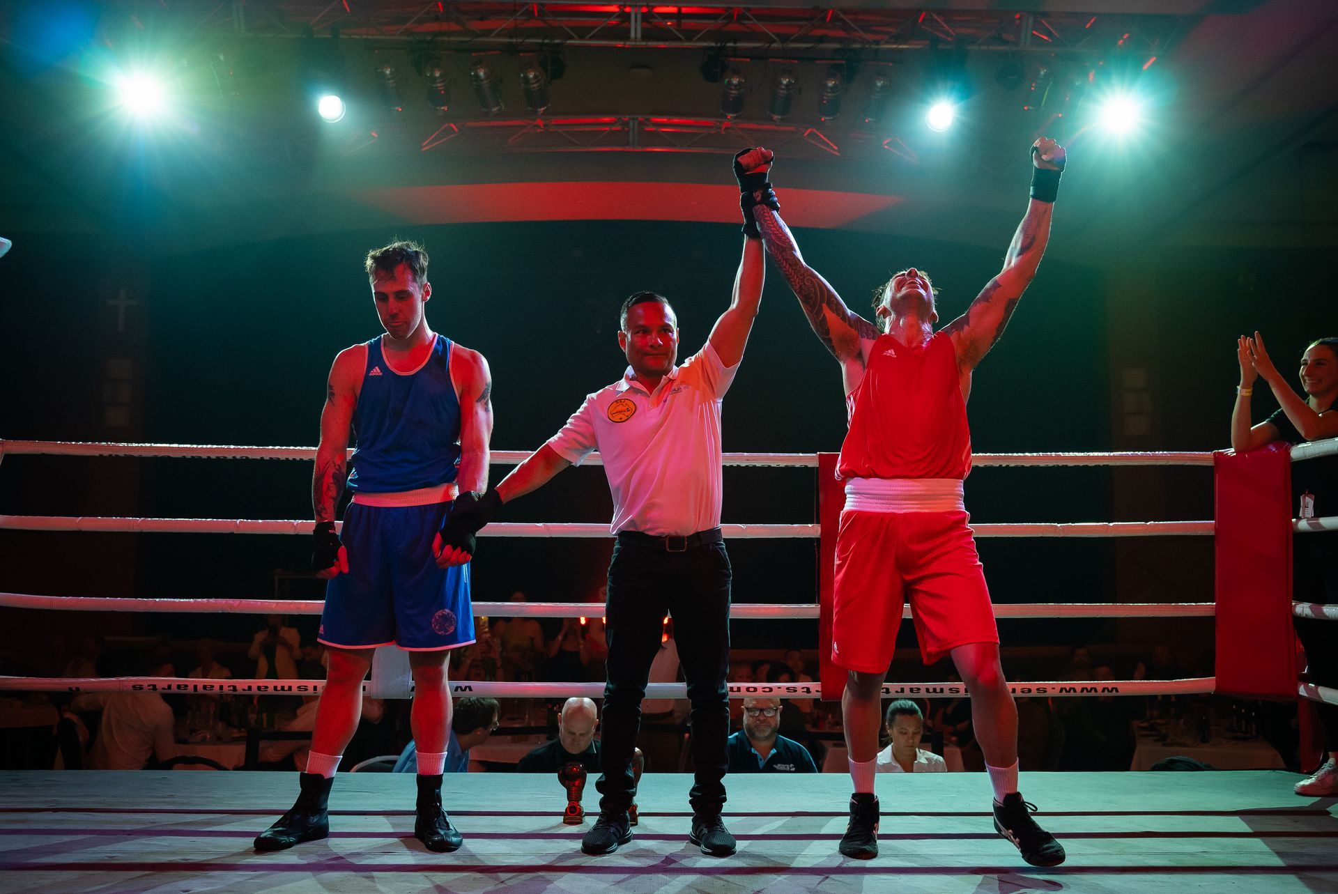 Boxer in red wins match, arms raised. Referee and defeated boxer in blue stand in ring.