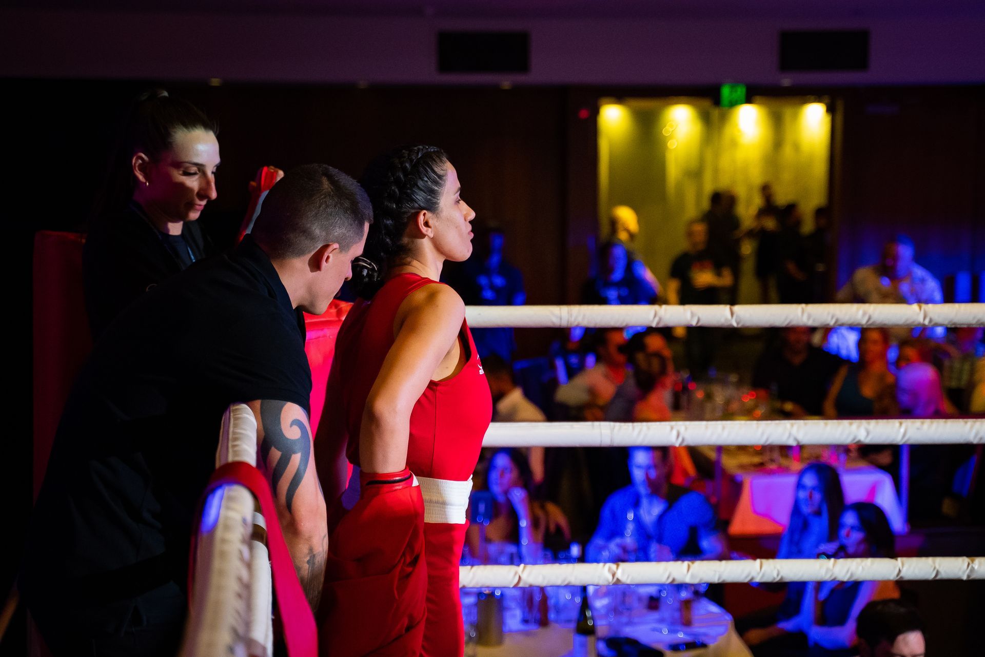 Boxer in red gear in the ring, flanked by a coach and another person, with an audience in the background.