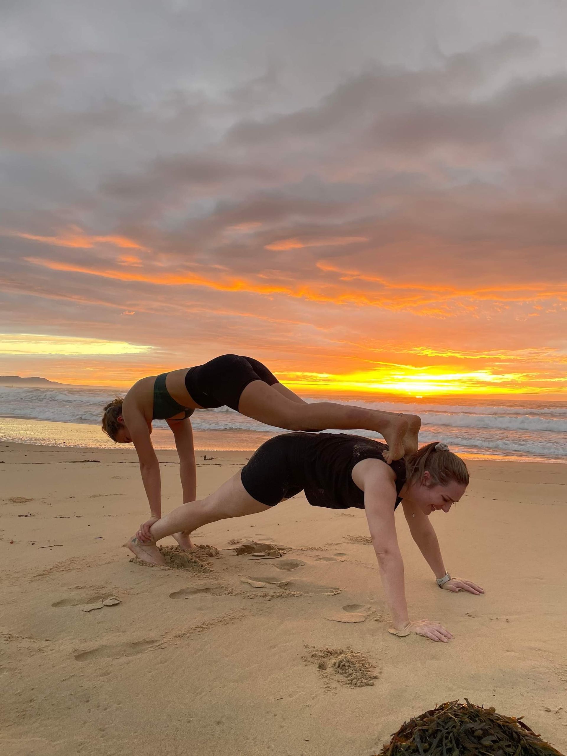 Two people doing a plank pose on a beach at sunset, one person stacked on top of the other.