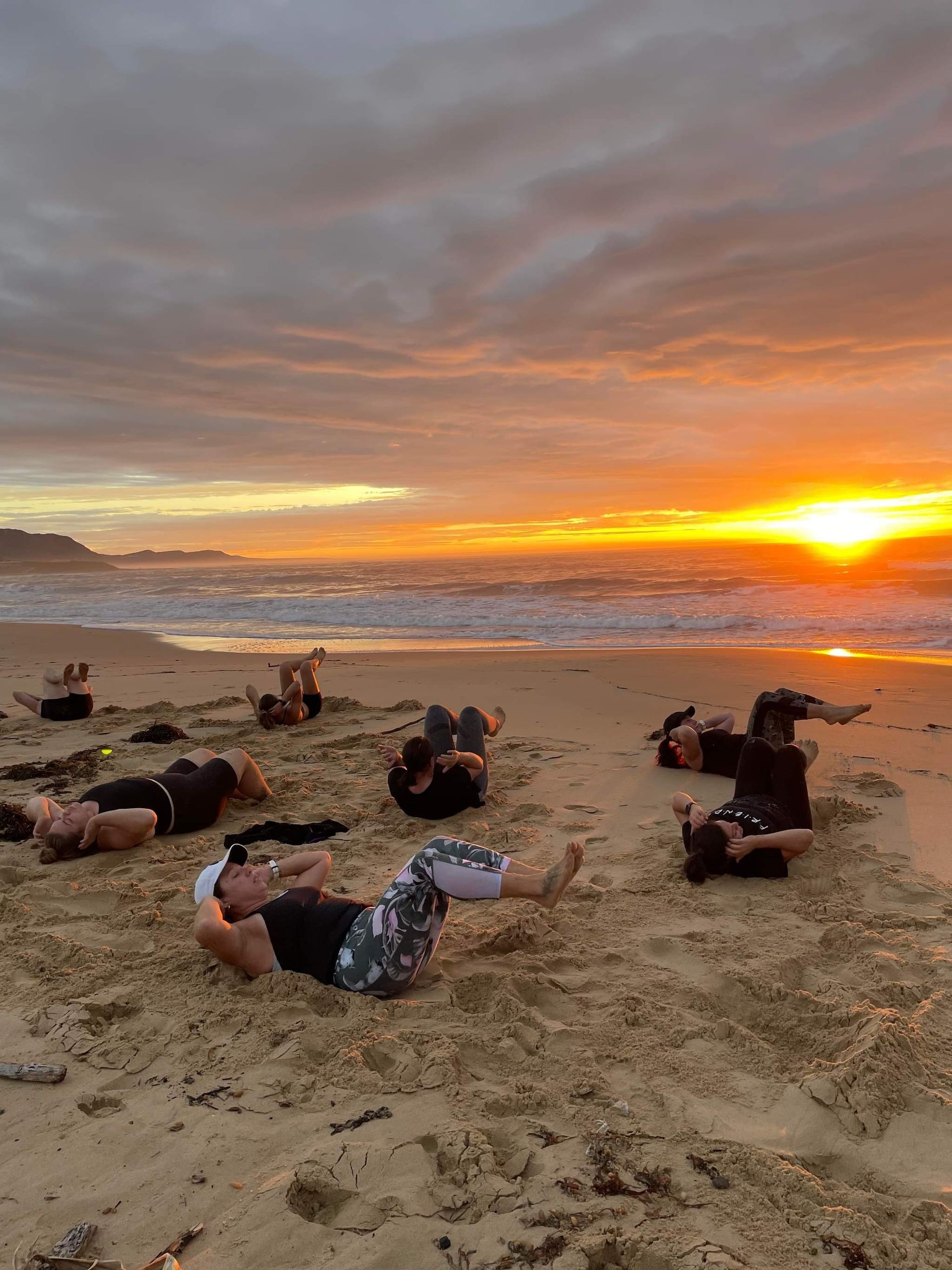 People exercising on a sandy beach at sunrise; ocean waves in background; orange sky.