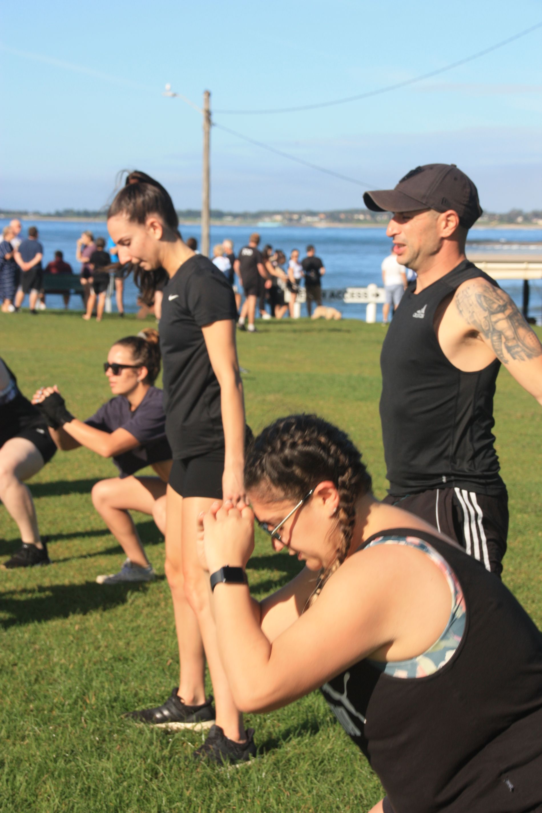 People doing squats on grass near water. A man in a cap watches.