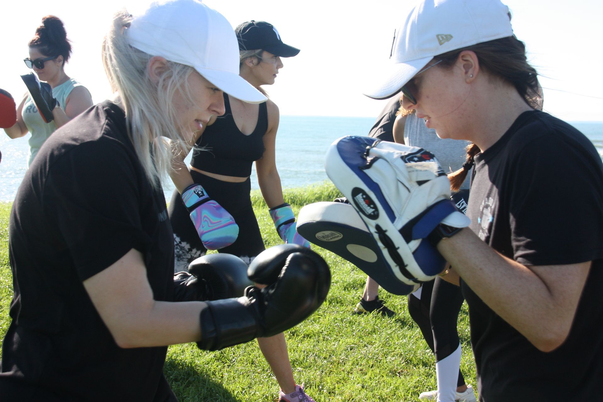 Two women boxing outdoors, one holding pads, the other wearing gloves.