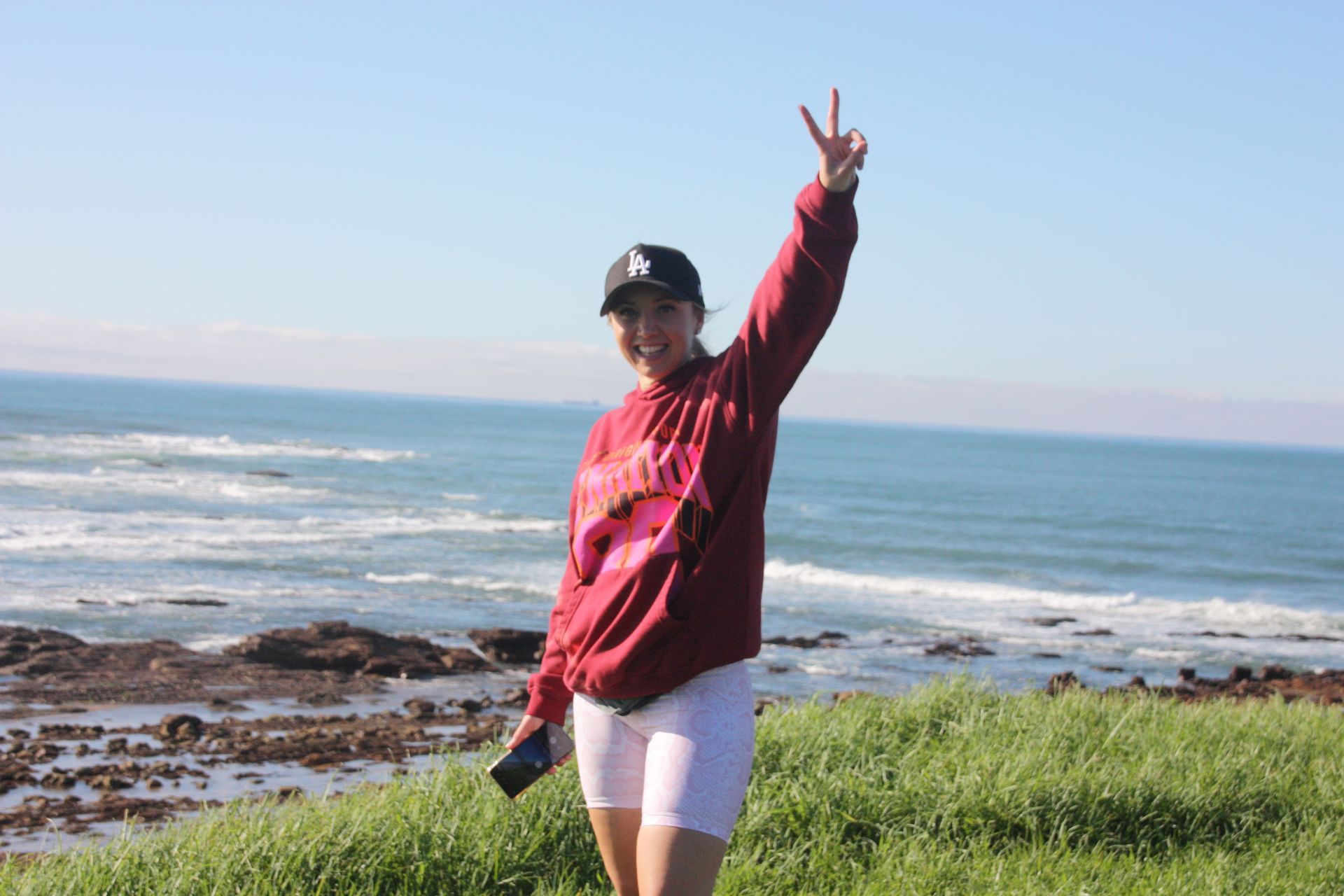 Woman with peace sign by the ocean, wearing a maroon hoodie, white shorts, and a baseball cap.