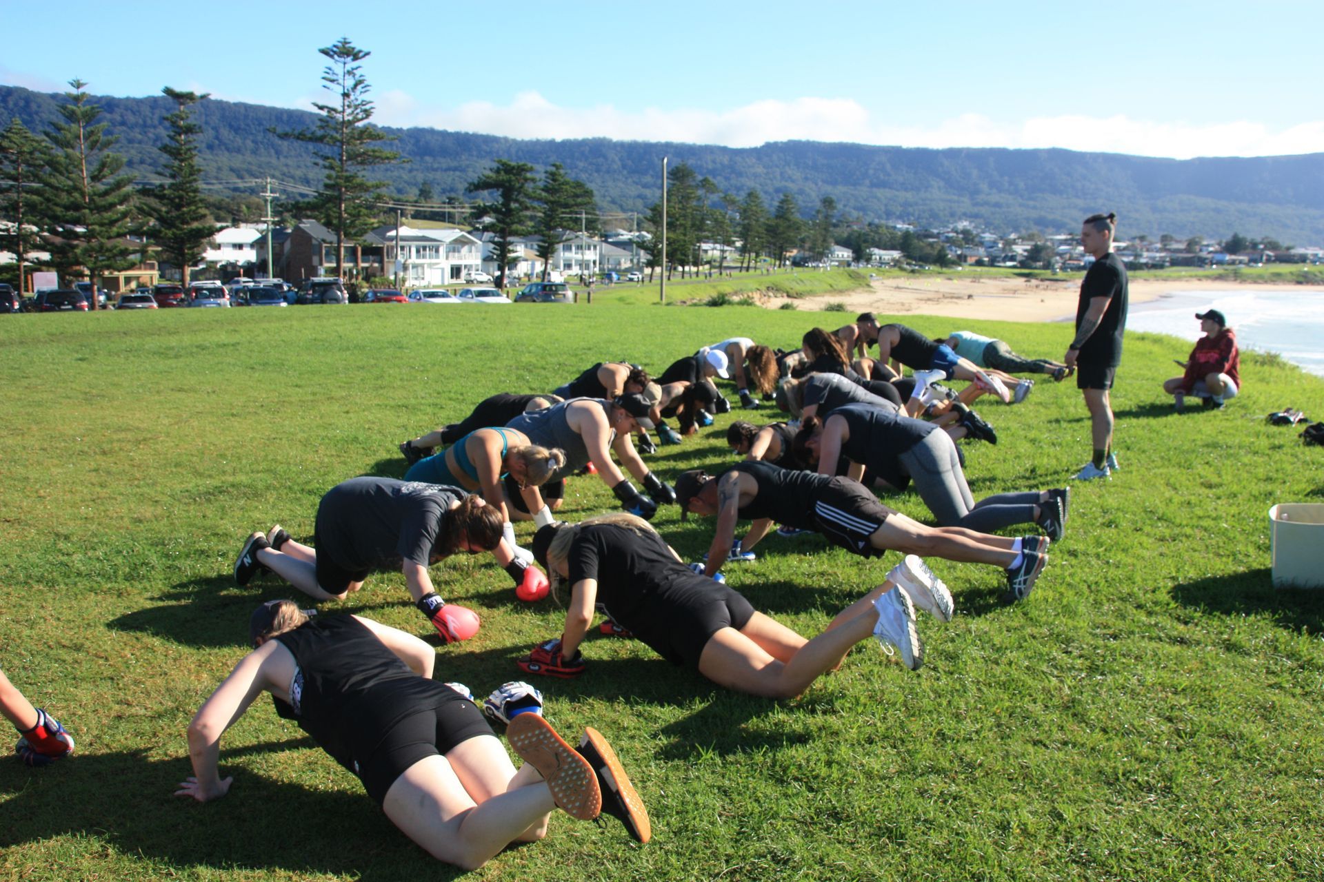 A group of people doing push-ups on grass by a beach. A man supervises them.