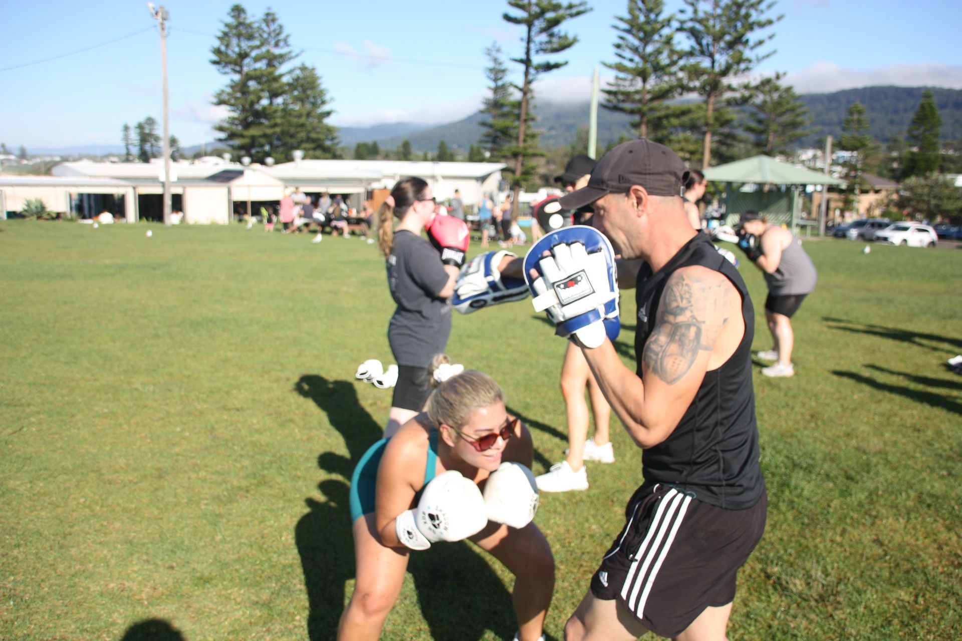 People in a grassy field training with boxing gloves; instructor working with a student.