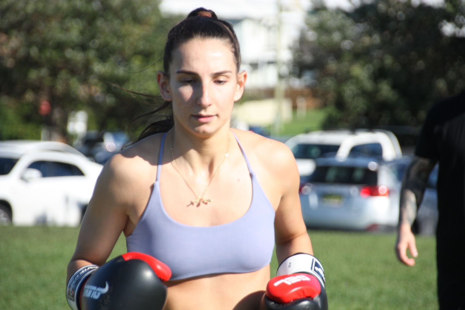 Woman in boxing gloves, working out in a park, wearing a sports bra, focused expression.