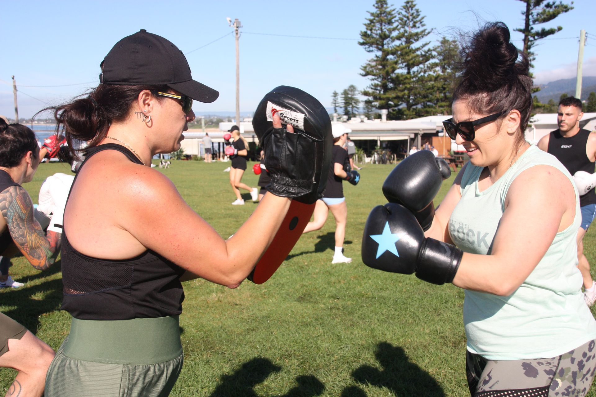 Two women boxing outdoors, one holding pads. Others in background.