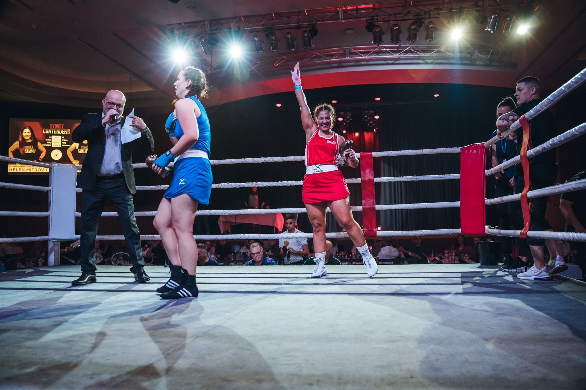 Boxer in red uniform celebrates victory in ring, announcer with microphone, blue-uniformed boxer looks on.