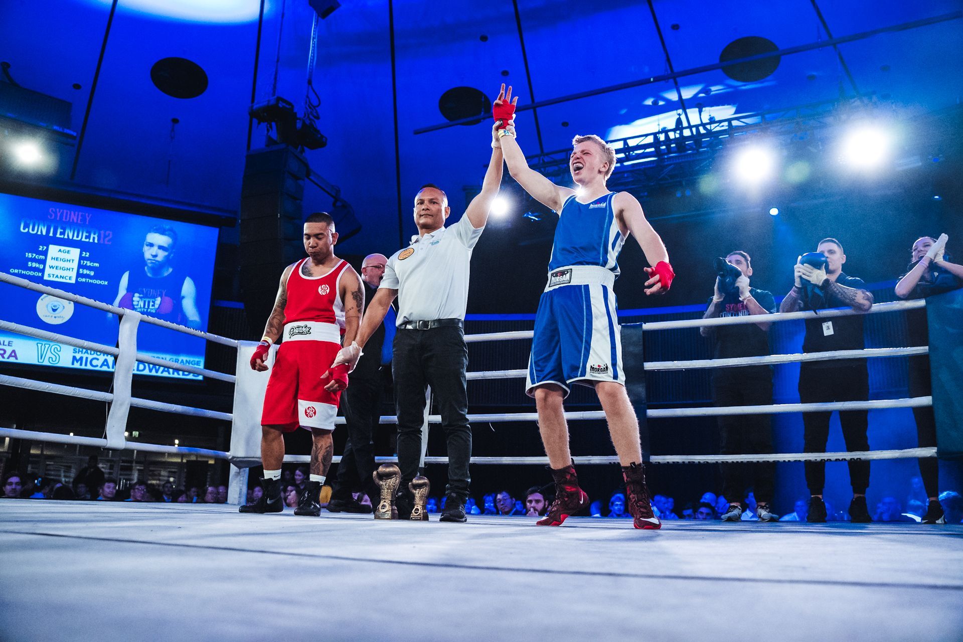 Boxer in blue raises arm in victory, referee raises his hand; opponent in red stands nearby on boxing ring.