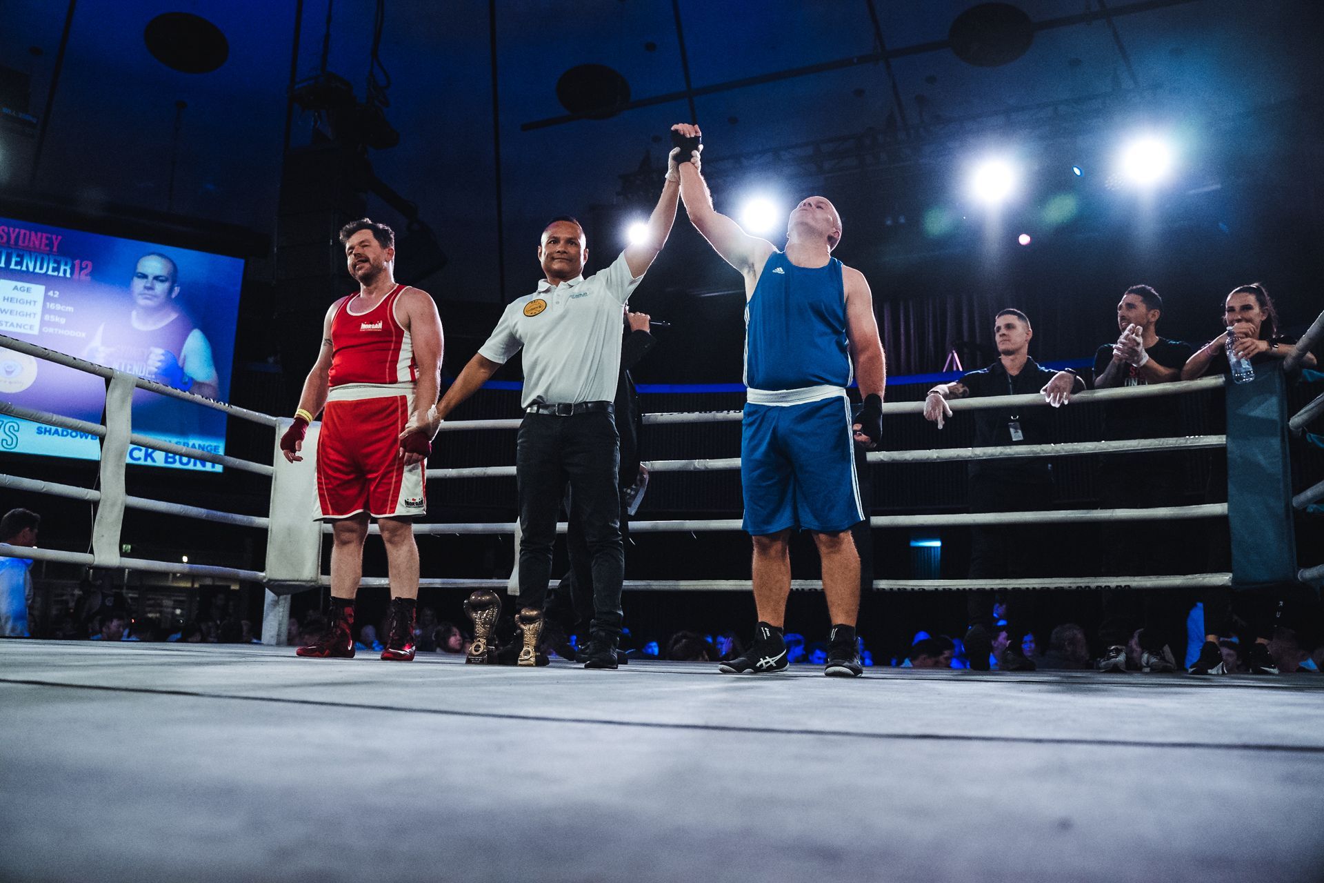 Boxer in blue shorts and tank top has arm raised, declared the winner in boxing ring.