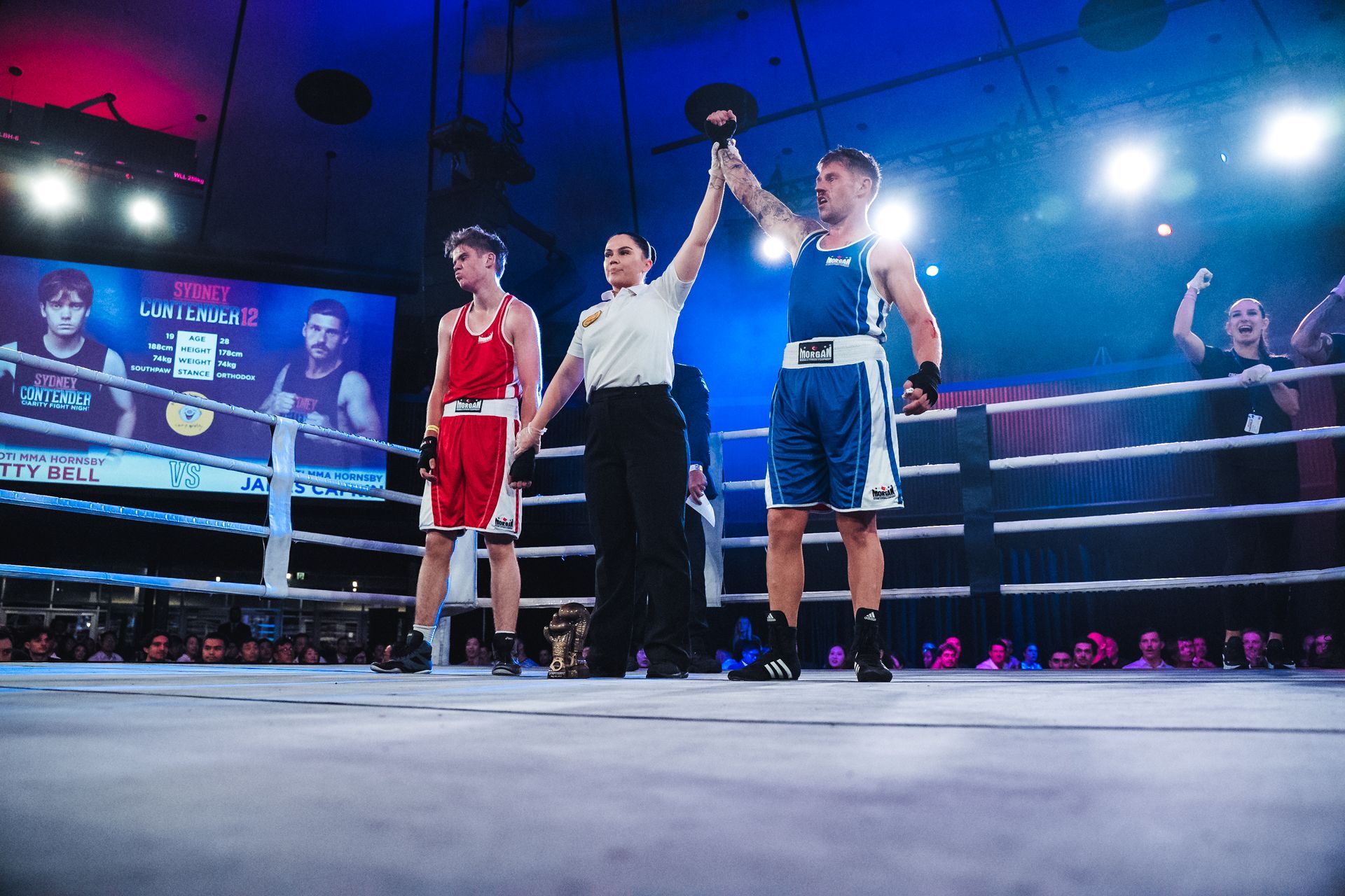 Boxer in blue uniform wins match in a boxing ring, referee raises his arm. Opponent in red looks on.