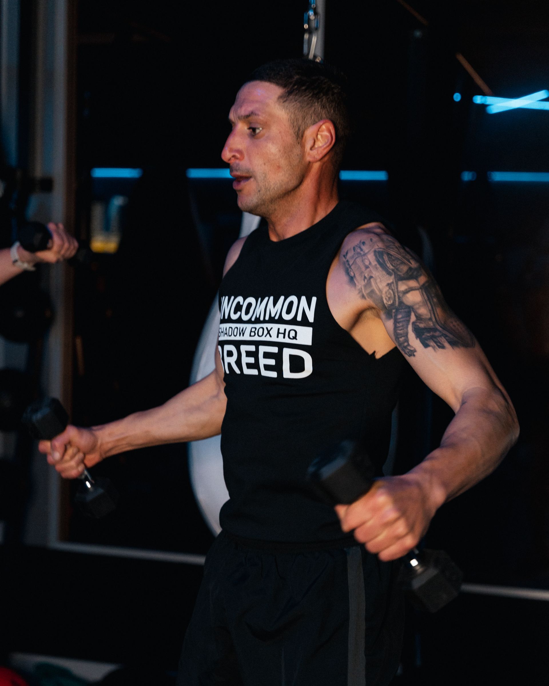 Man in black tank top exercising with dumbbells in a gym setting.