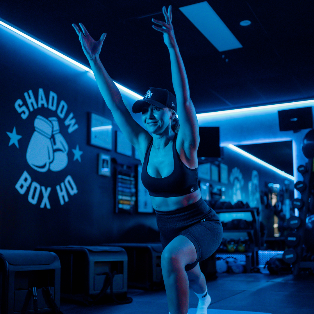 Woman in athletic wear, arms raised, practicing yoga in blue-lit gym. Shadow Box HQ logo visible.