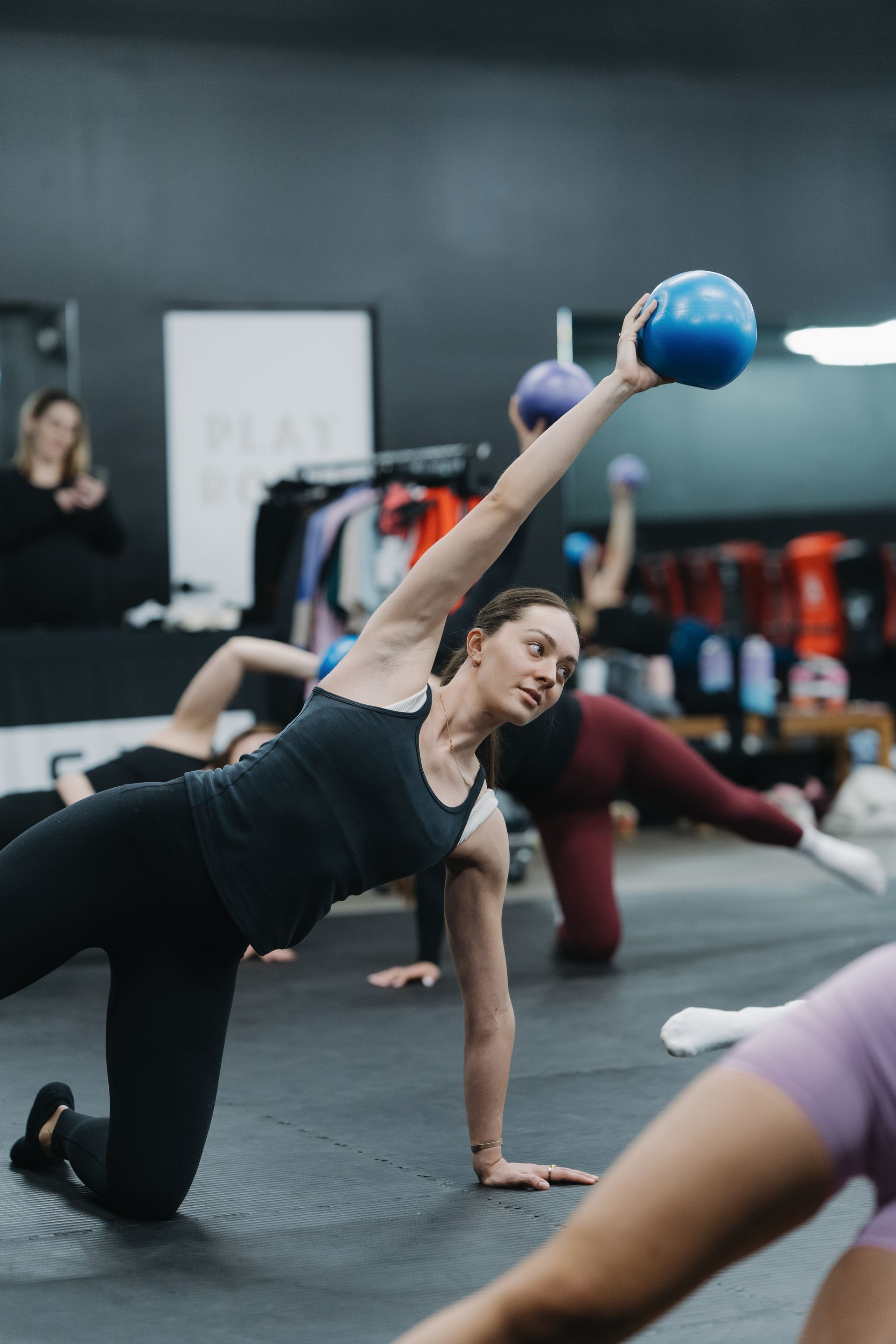 Woman in a fitness class kneels, holding a blue ball overhead. Other people exercise in a studio.