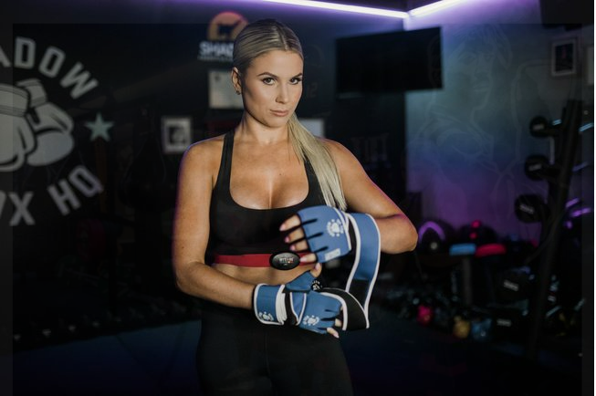 Woman in gym attire putting on boxing gloves, serious expression. Shadow Box HQ logo in background.