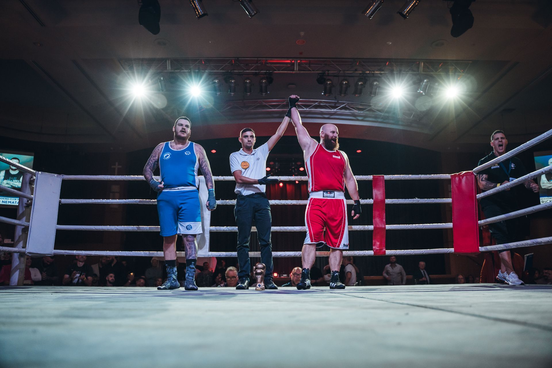 Boxer in red with raised arm is declared winner in boxing ring, referee stands between him and opponent.