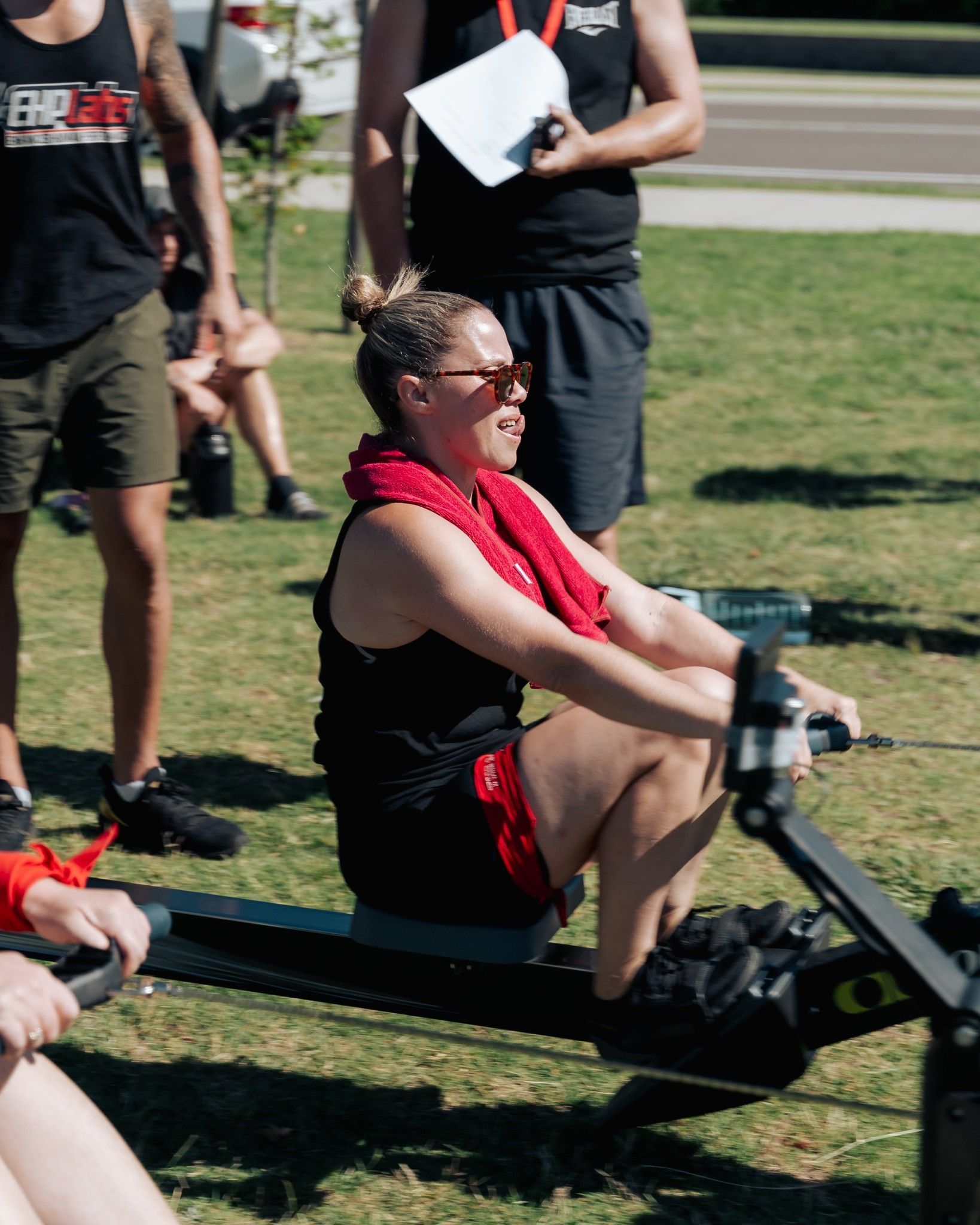 Woman rowing outdoors, wearing black athletic wear and sunglasses, red towel around neck.