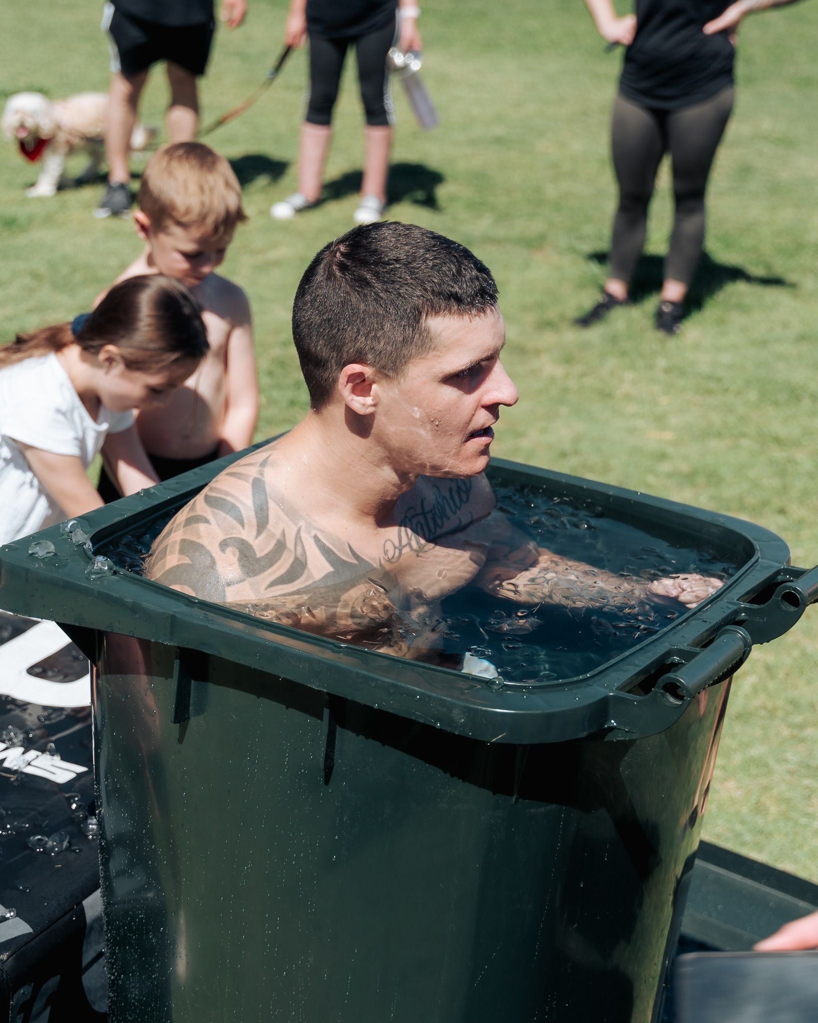 Man in a trash can filled with cold water, outside. Has tattoos on arms, surrounded by people in a park on a sunny day.