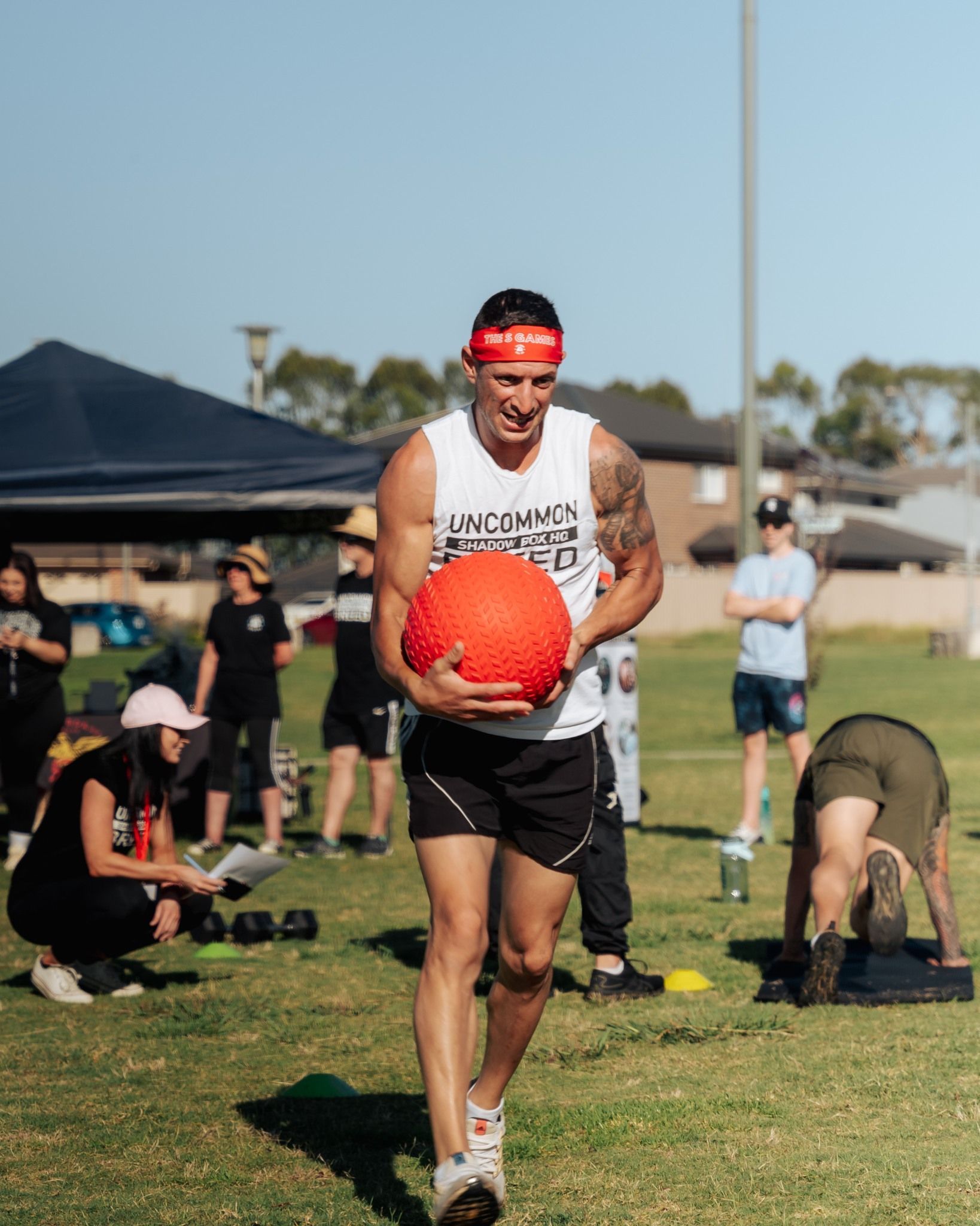 Man in headband and athletic wear carries a red ball on a grassy field; others exercise in the background.