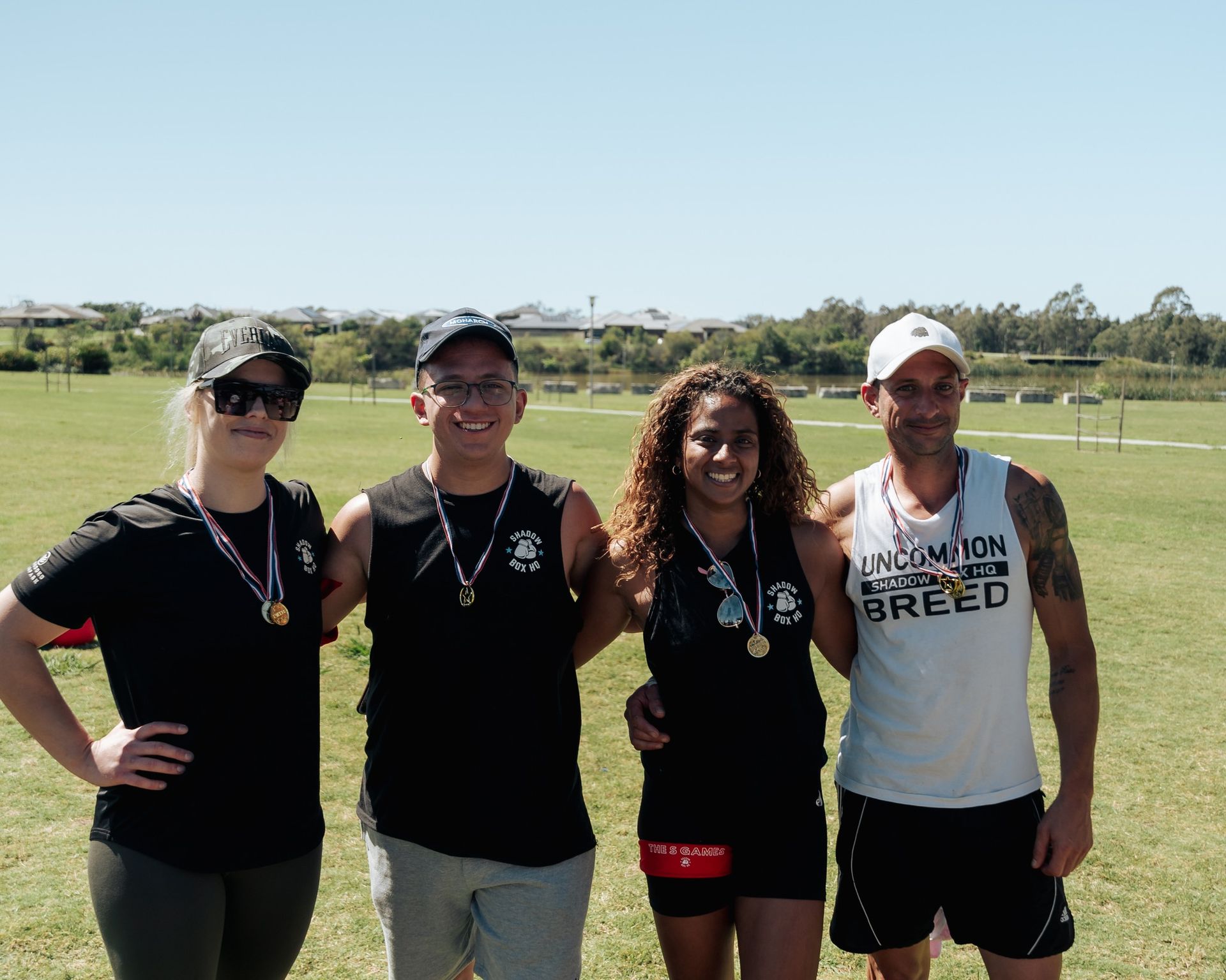 Four people with medals pose outdoors, smiling. They wear athletic clothes on a sunny day.