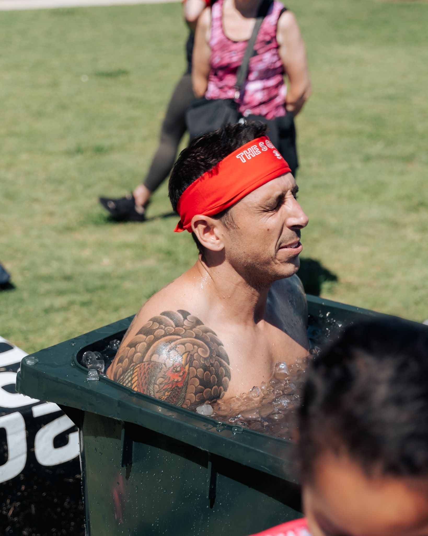 Man in a red headband, shirtless in a green tub of icy water, with a tattoo on his shoulder, in a park.