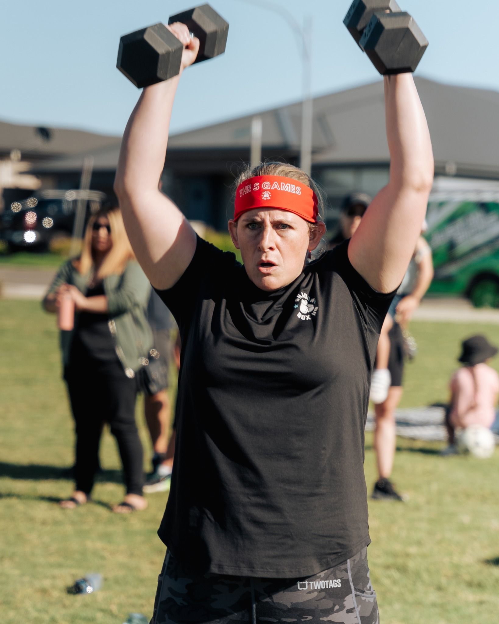 Woman in black shirt and camo pants lifts dumbbells overhead outdoors. Red headband.