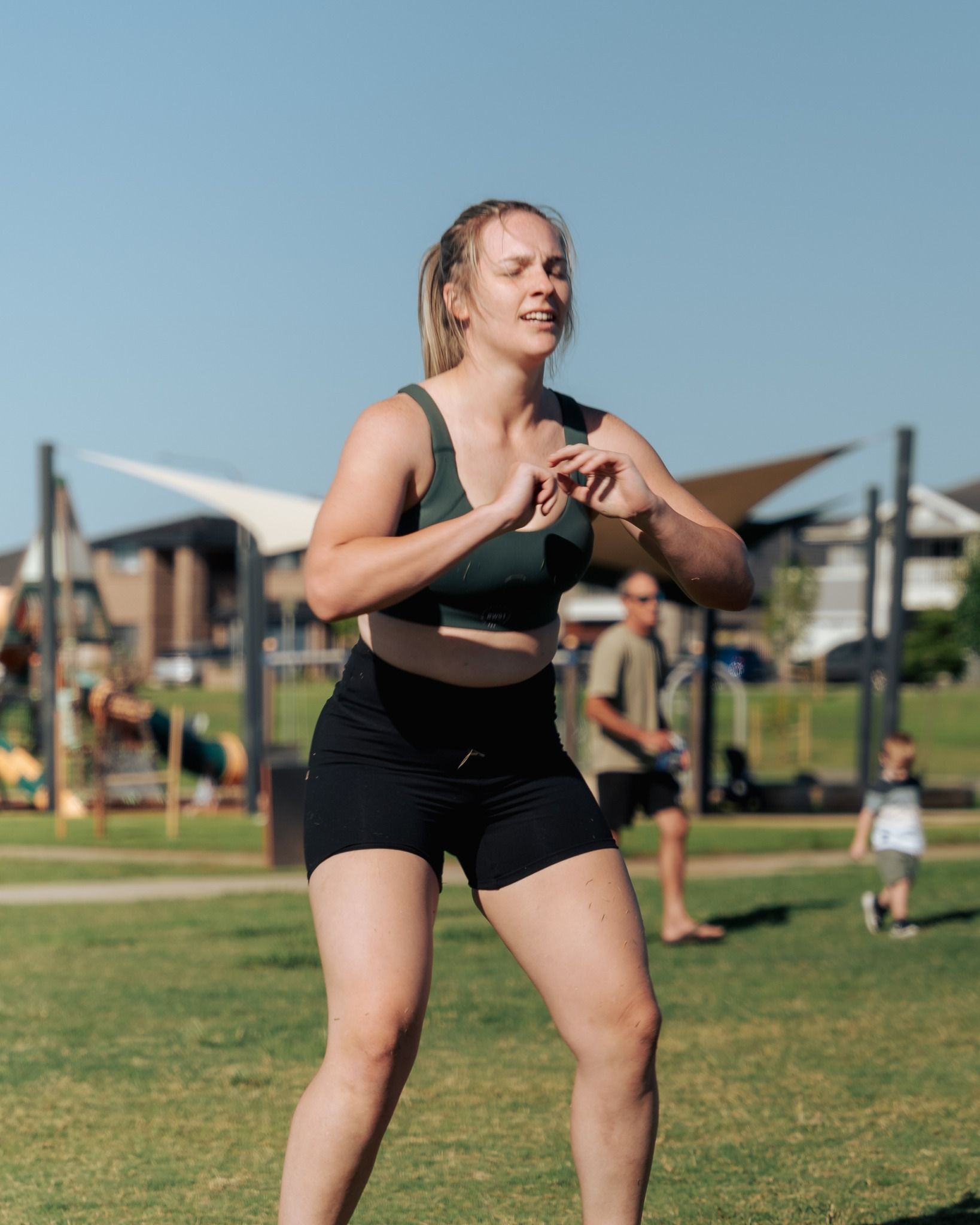 Woman exercising in a park, wearing a green sports bra and black shorts, squatting, sunny day.