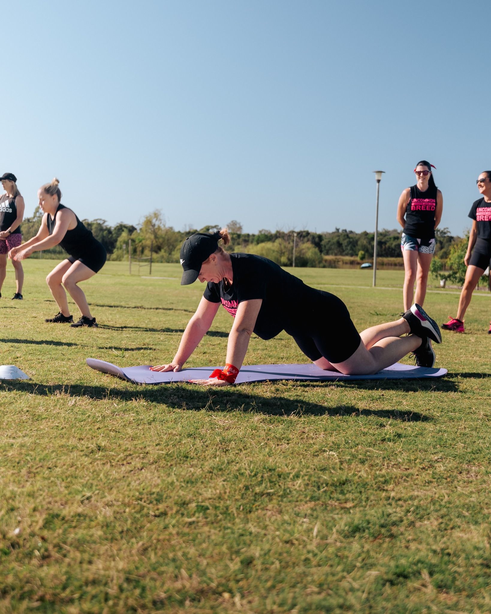 Group of people exercising outdoors; one does push-ups on a mat, others squat.