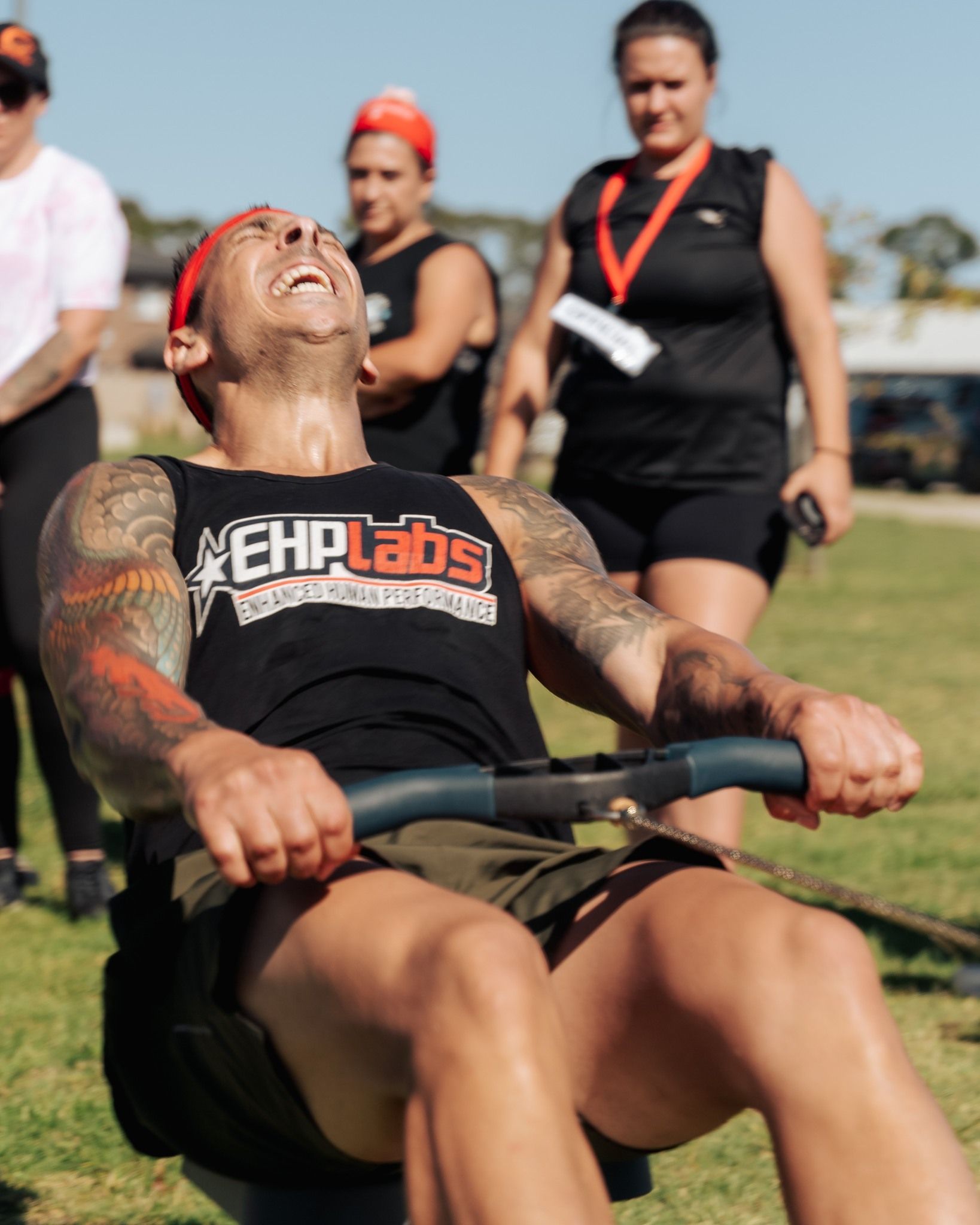 Man rowing a machine outdoors, face contorted in effort, wearing an EHP Labs tank top; others watch.
