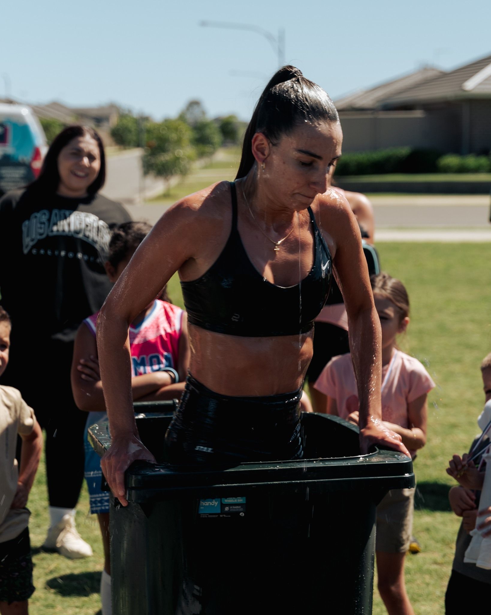Woman in sports bra and shorts sits in a trash can of water, surrounded by children and a woman on a sunny day.