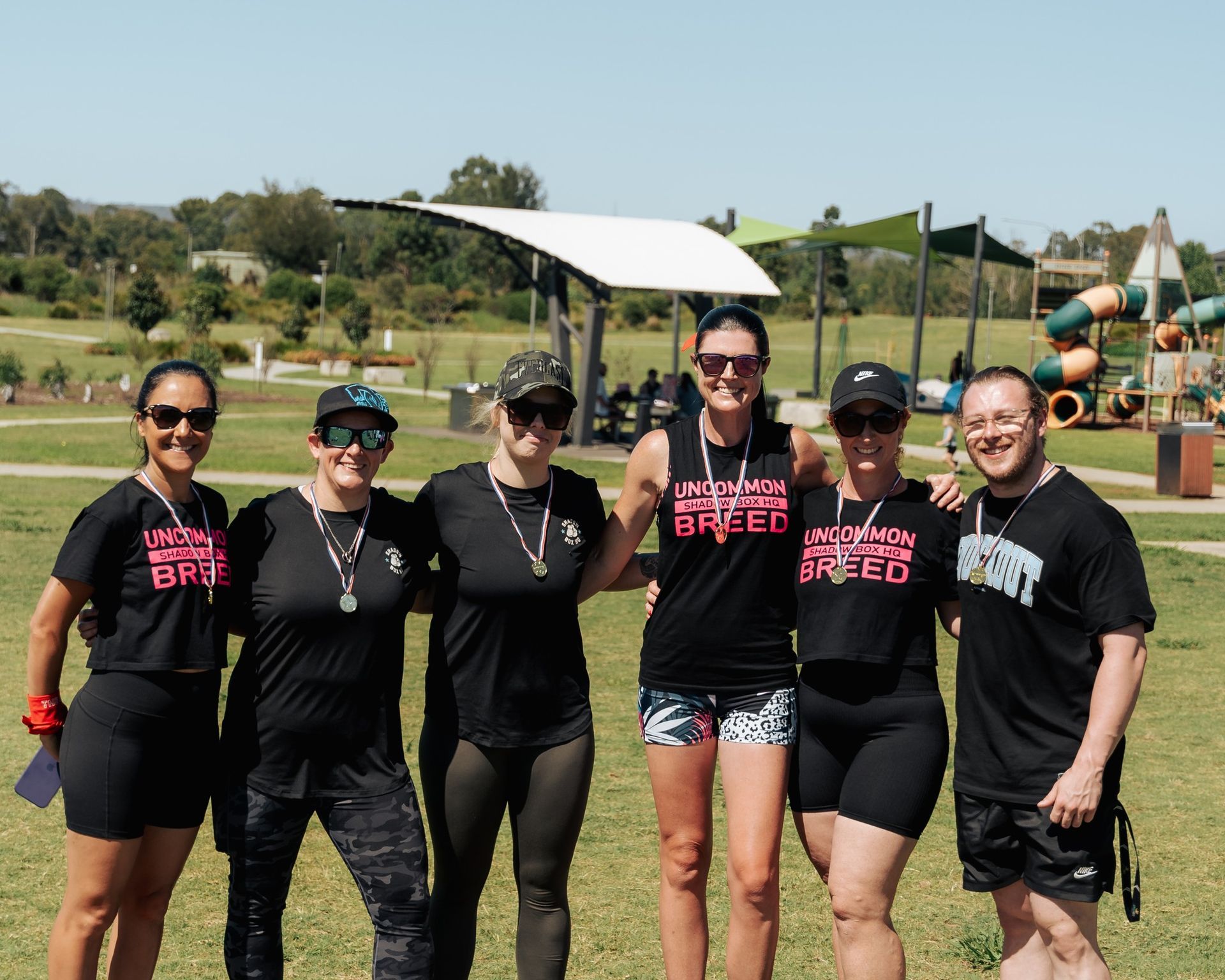 Group of six people posing together outdoors. They wear athletic gear, some with medals. Park setting.