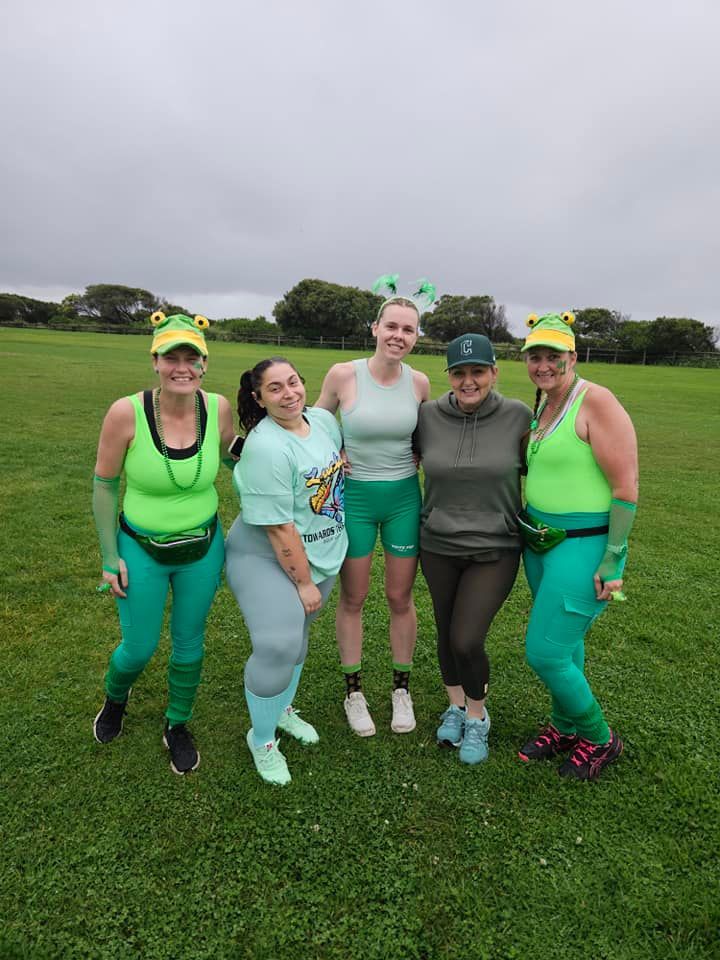 Five people pose on a grassy field, most in green outfits with festive headbands. Overcast sky.