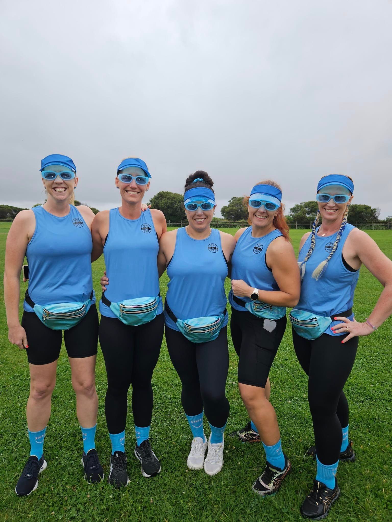 Five people in blue outfits with sunglasses and fanny packs stand on a grassy field.