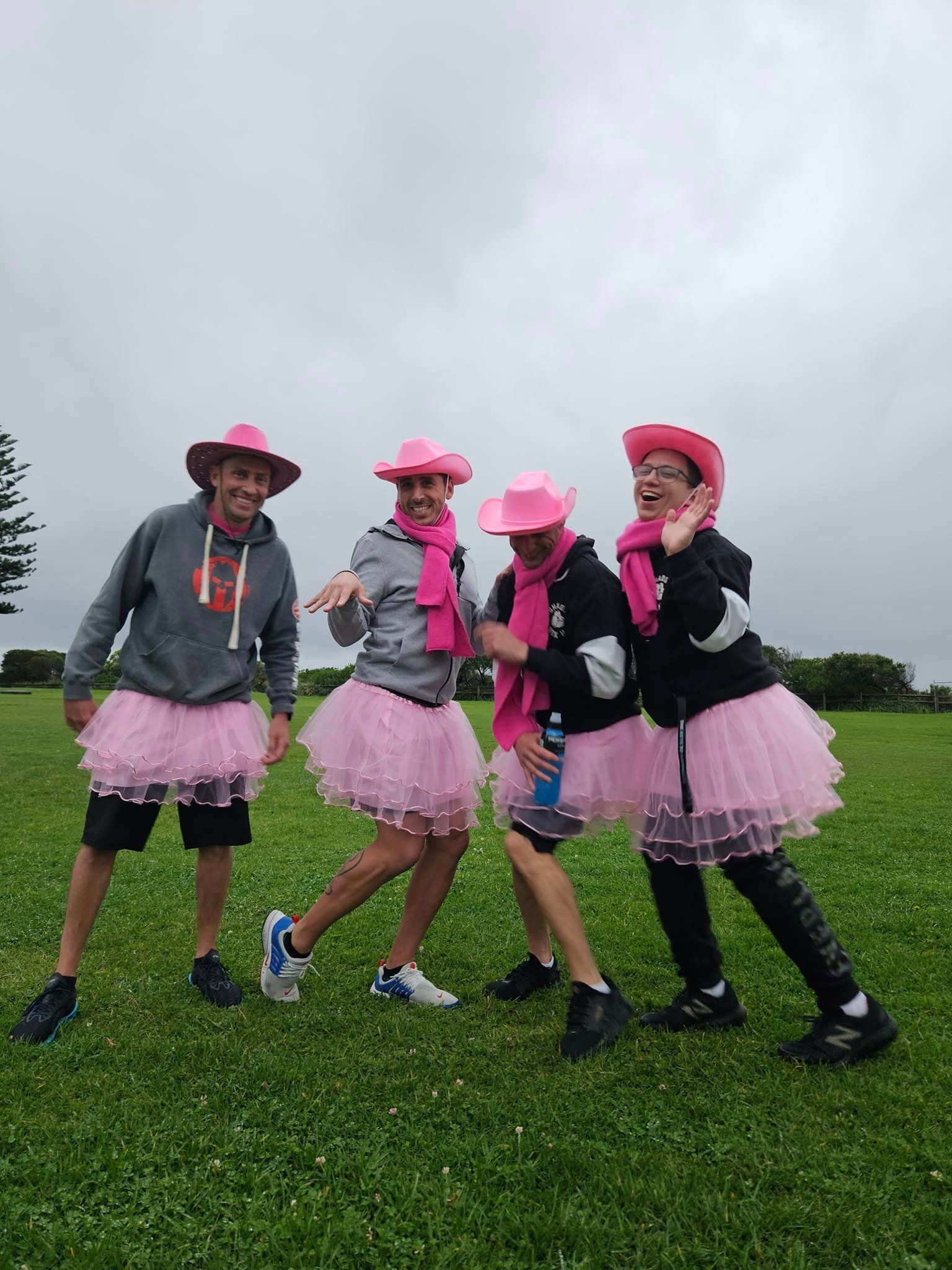 Four people in pink cowboy hats, tutus, and scarves, posing on a grassy field under a cloudy sky.