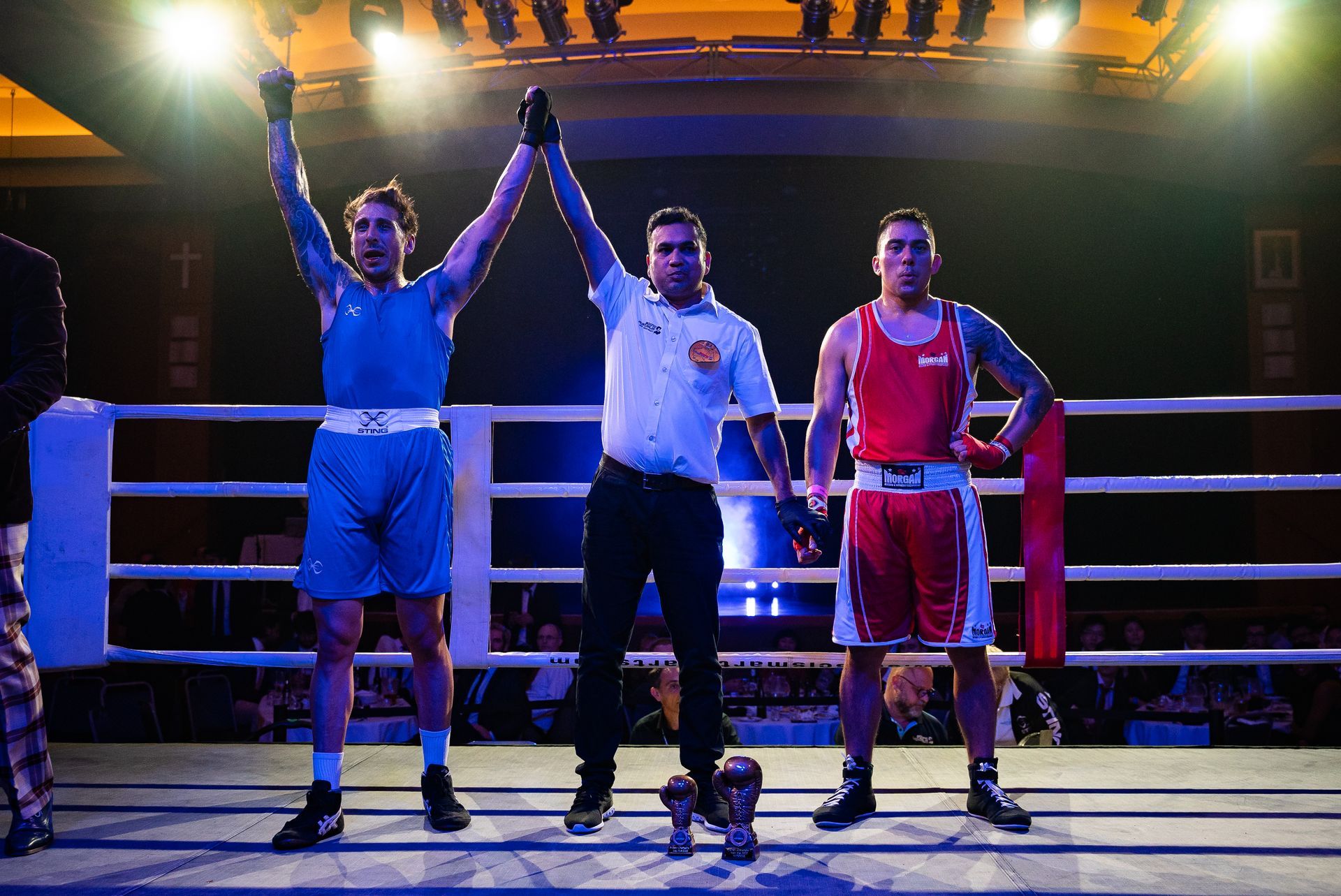 Boxer in blue wins; referee raises their arm. Red-clad boxer stands, ring is lit with bright lights.
