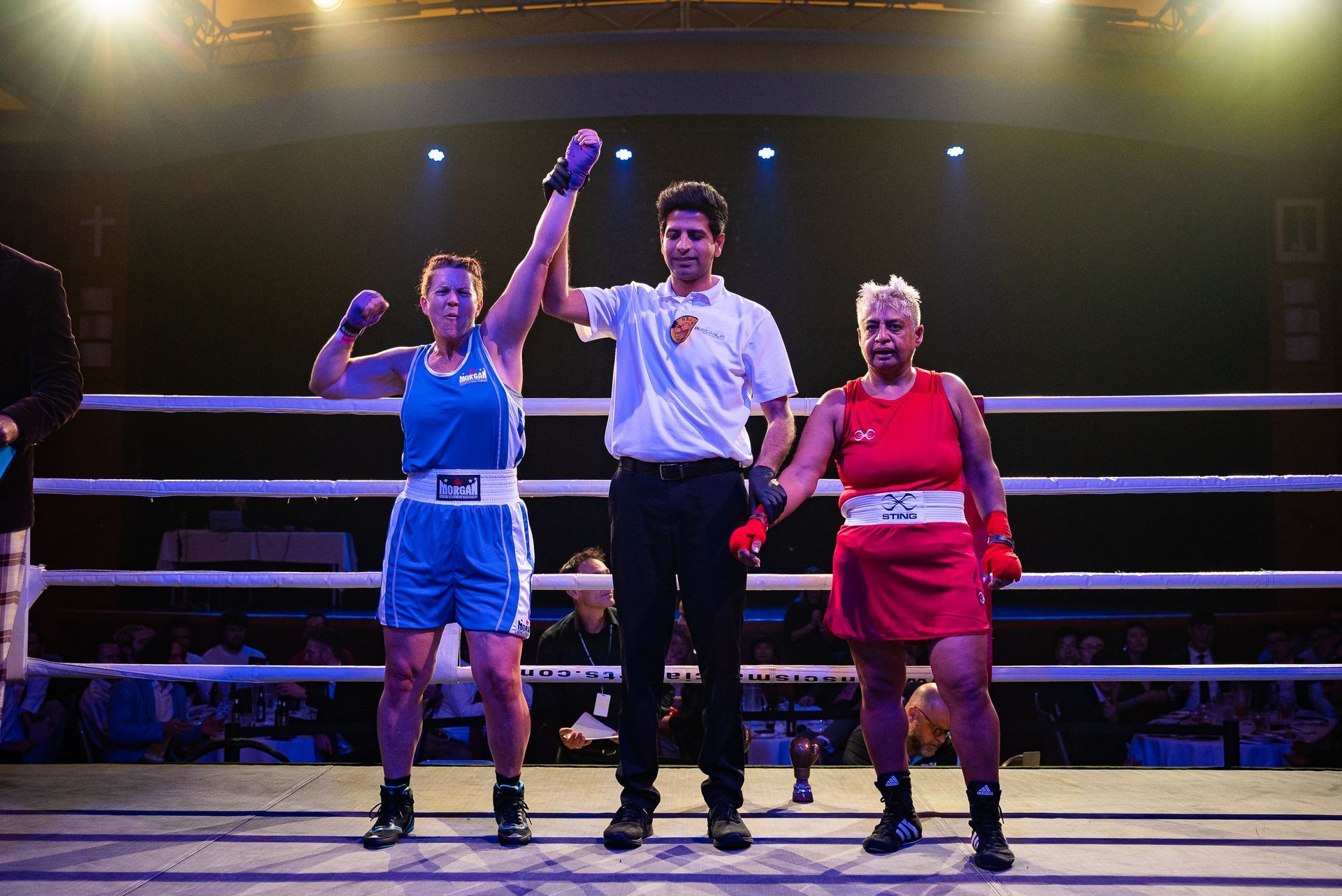 Boxer in blue raises arm after victory, referee gestures, other boxer in red, boxing ring.
