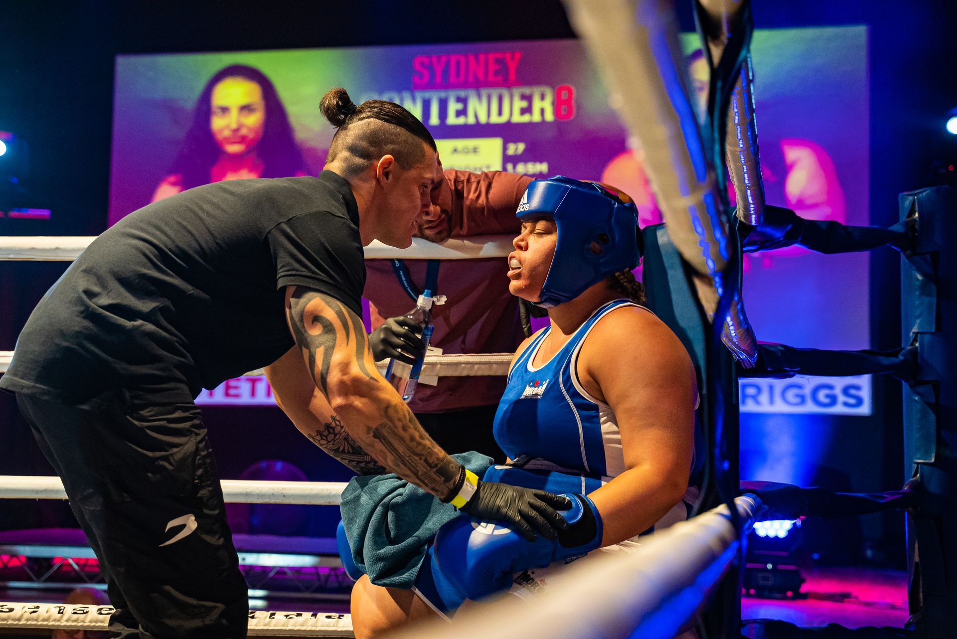 Boxer in blue protective gear receives instructions from coach in a boxing ring.