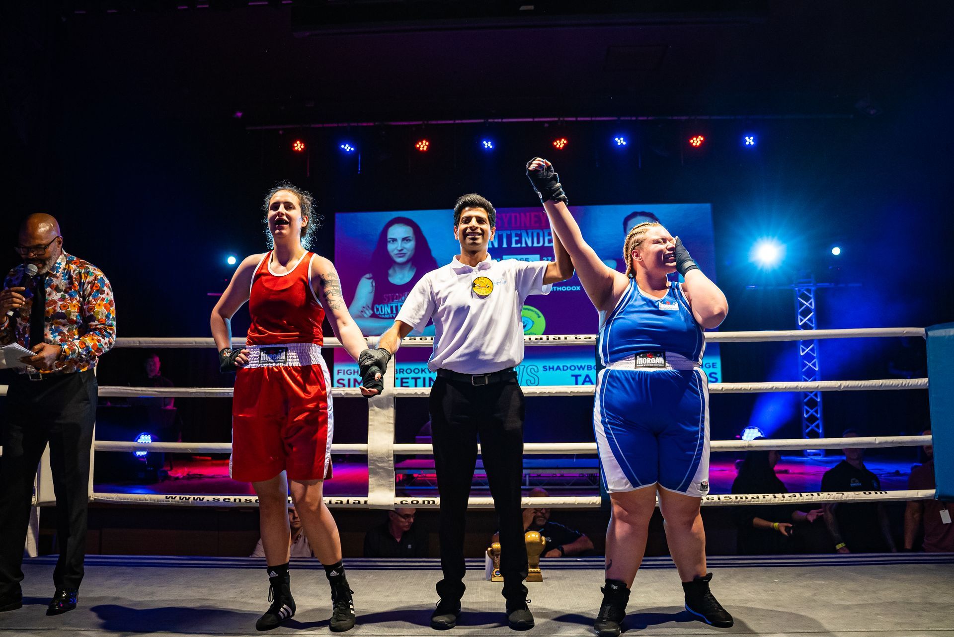 Two boxers in a ring after a match, one in blue raised in victory, referee between them, announcer at left.