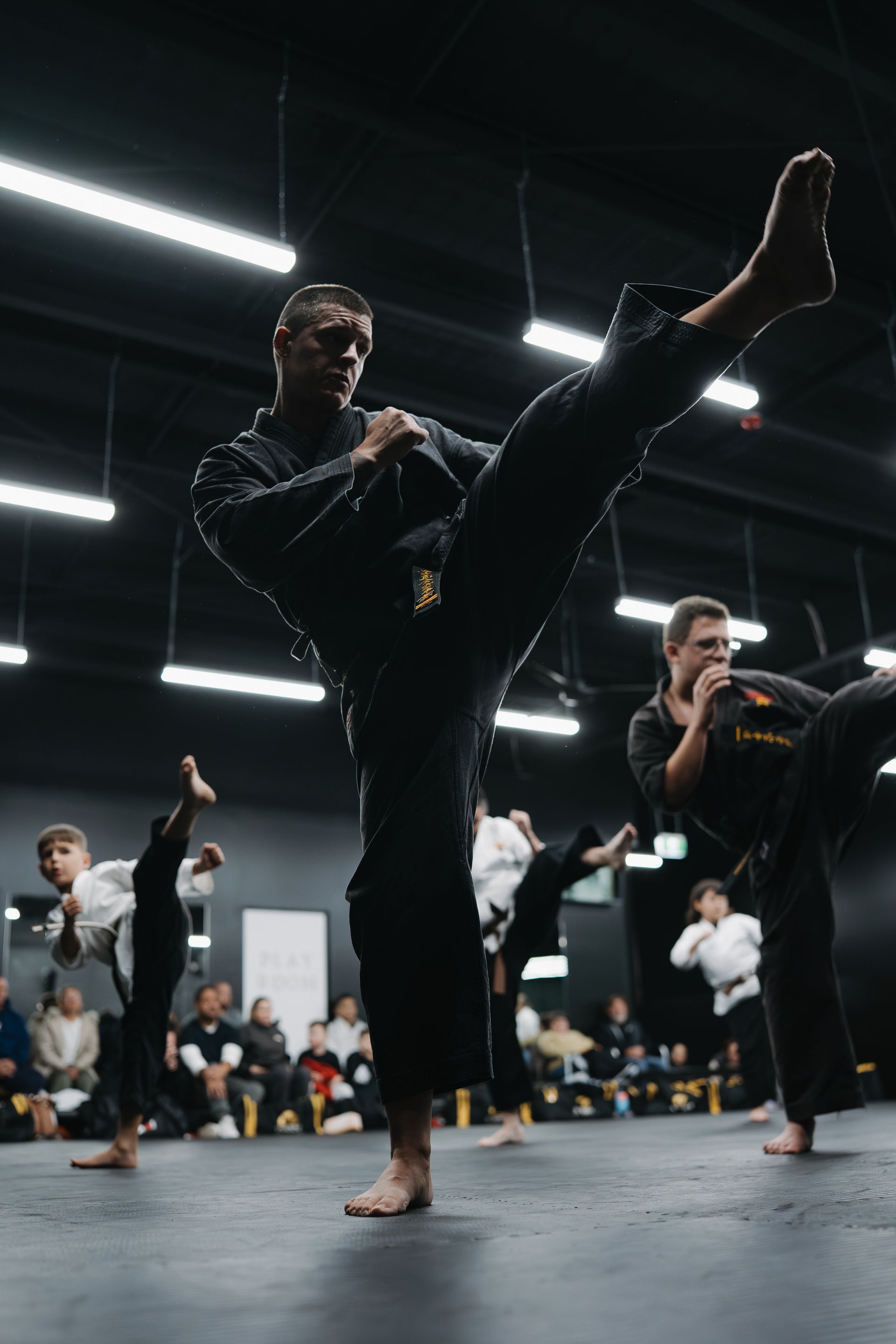 People in black uniforms practicing martial arts kicks in a dimly lit studio.